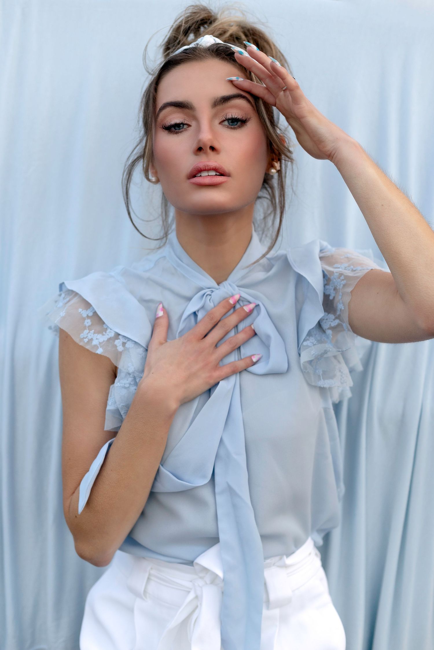 Fashion portrait of a model in a light blue ruffled blouse with white pants against a pale blue background.
