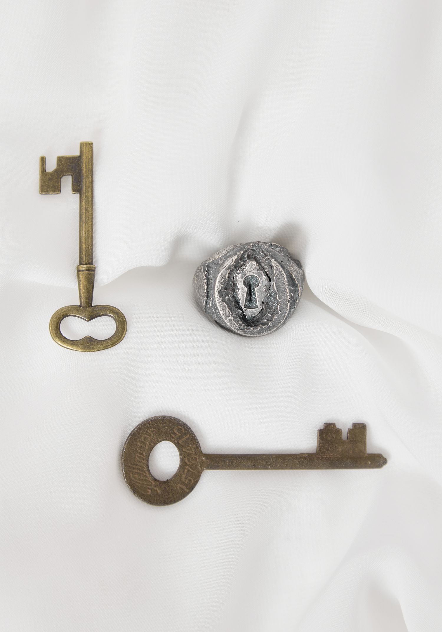 Two antique brass skeleton keys and a silver keyhole escutcheon arranged on white fabric.