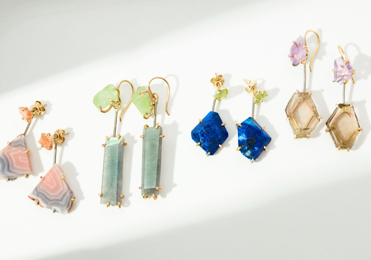 Collection of elegant gemstone drop earrings in blue, green, pink, and neutral tones with gold settings on white background.