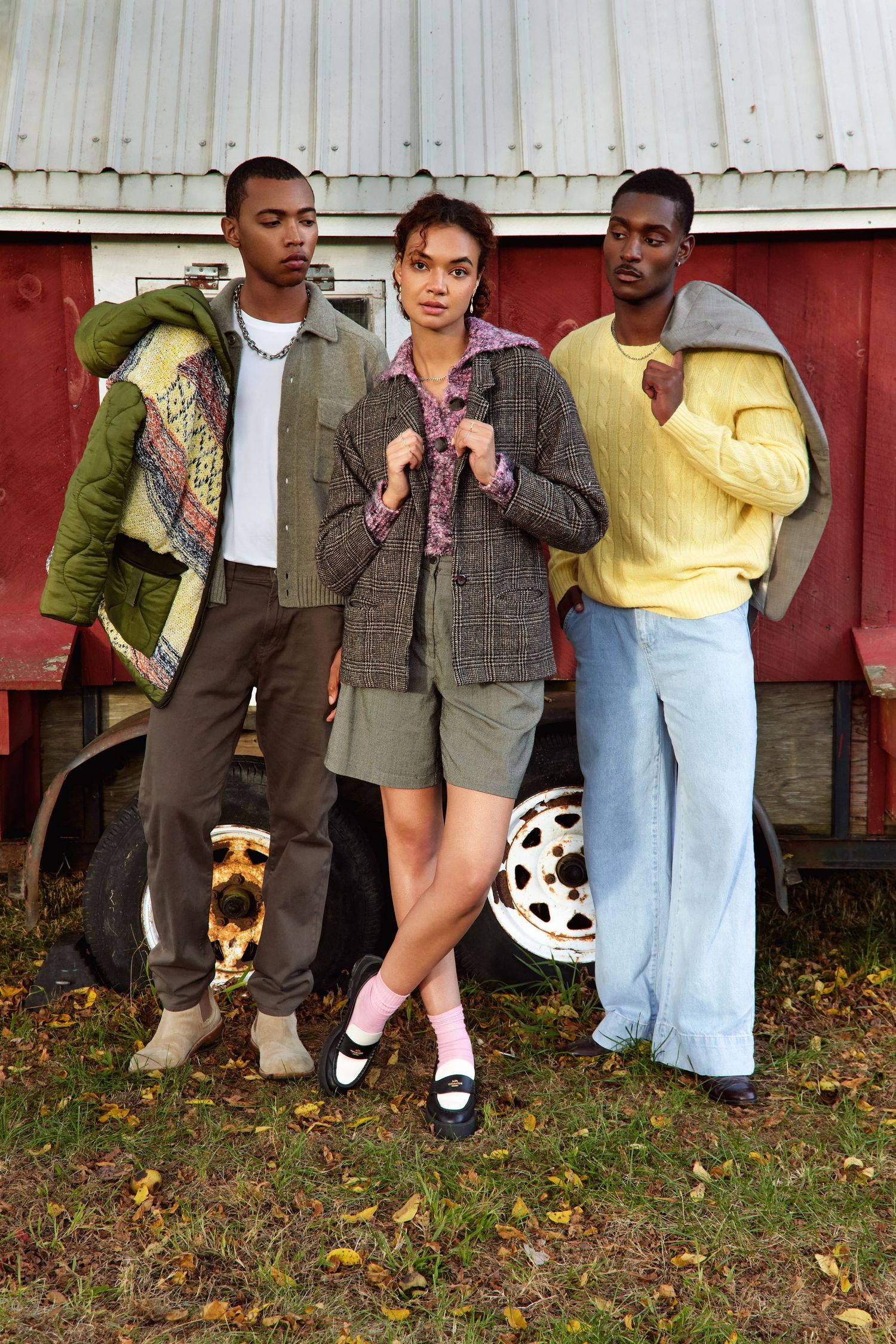 Three friends in trendy vintage-inspired outfits pose casually against a red barn wall in an autumn setting.