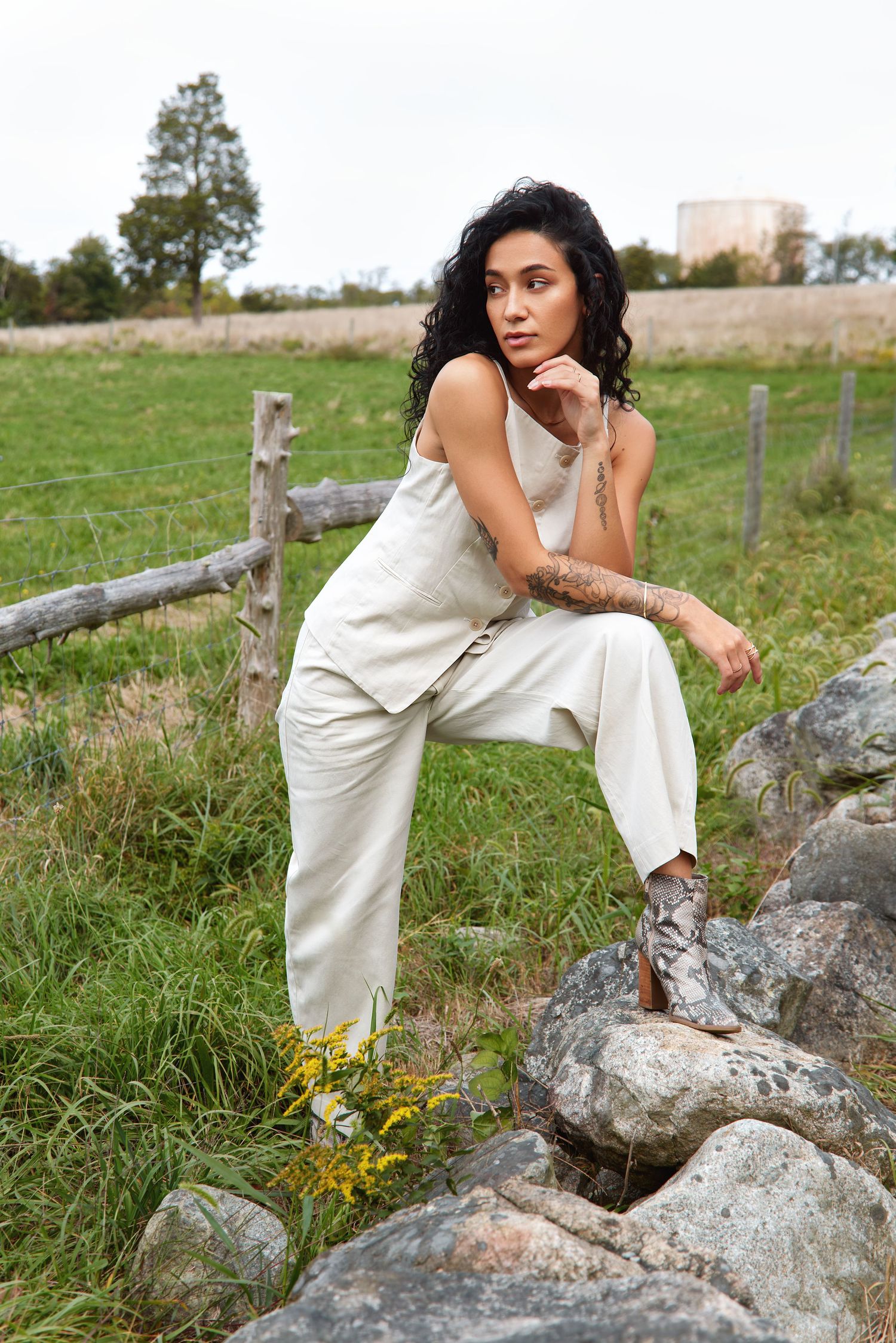 Person in white outfit poses thoughtfully on rocky terrain surrounded by green grass and wildflowers in a rustic setting.
