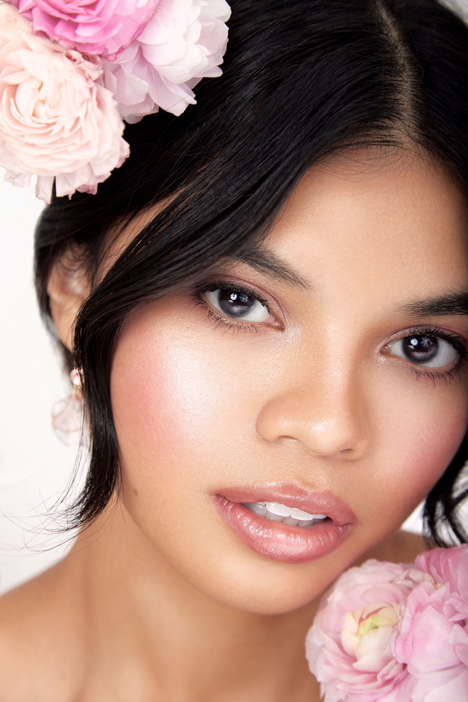 Soft pink roses frame a beauty portrait with natural glowing makeup and glossy lips against a light background.