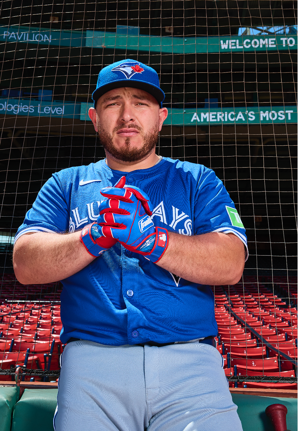 Baseball player in Toronto Blue Jays uniform sits in dugout at Fenway Park with red stadium seats visible in background.