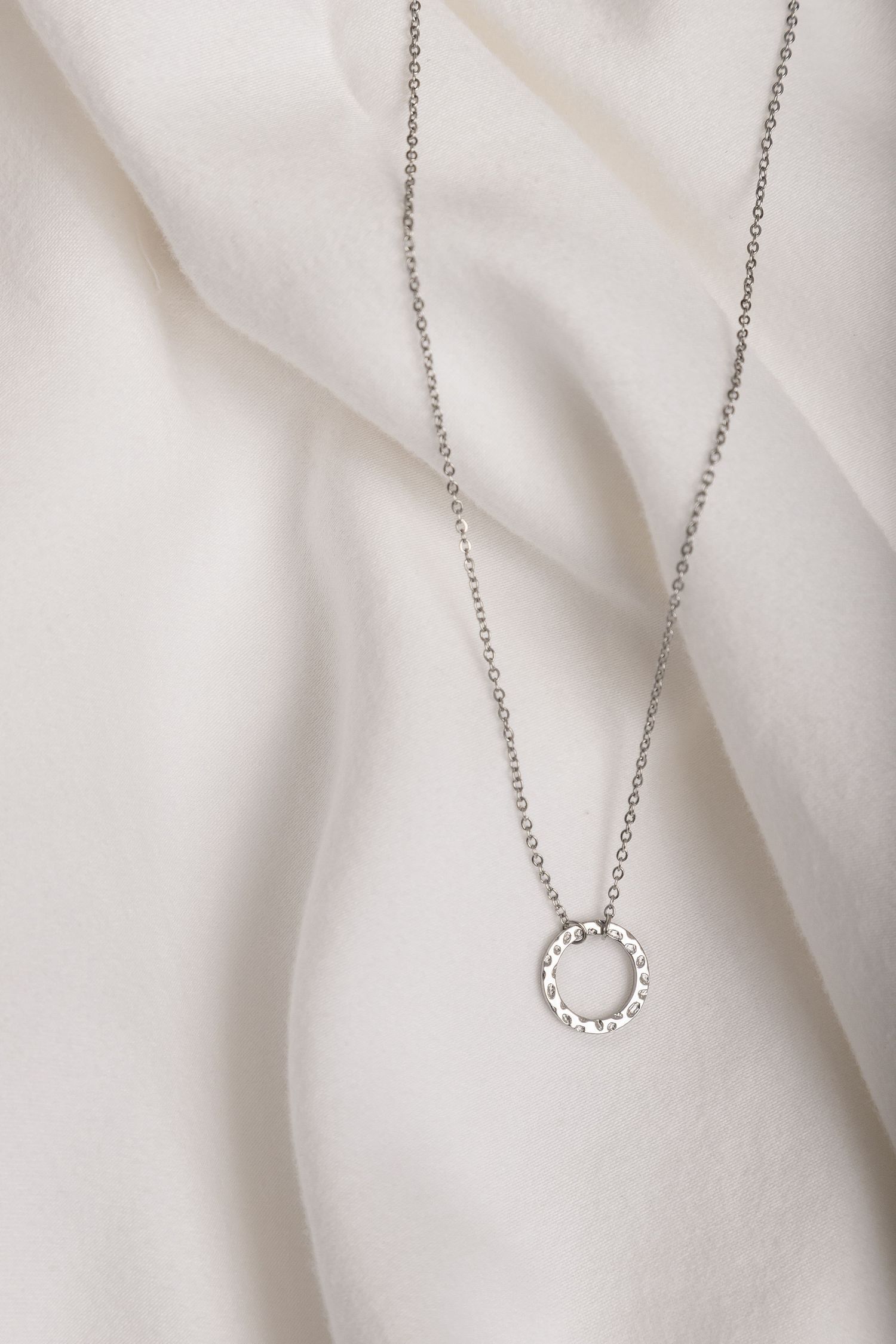 Delicate silver necklace with circular pendant displayed on soft white fabric with gentle folds and draping.