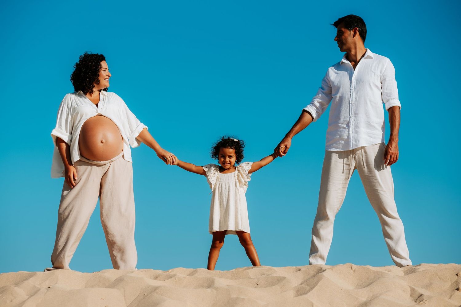 Family wearing white clothing holds hands while standing on a sandy beach against bright blue sky backdrop.