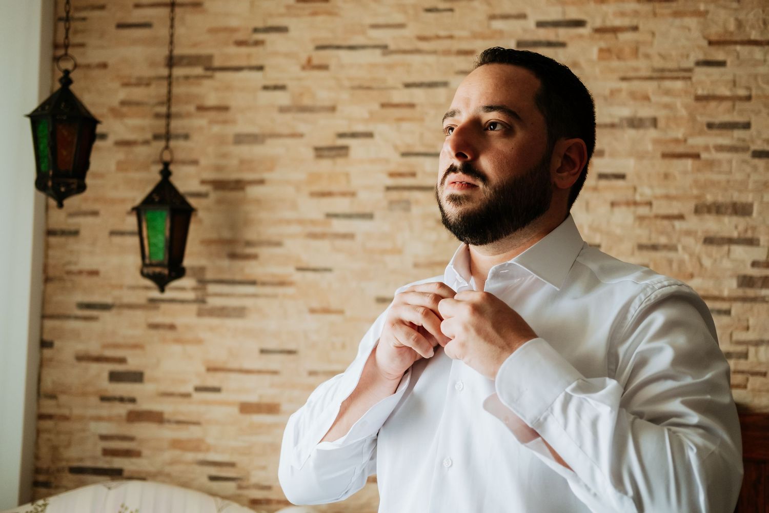 Groom in white dress shirt adjusts his cuffs while getting ready against a textured brick wall background.