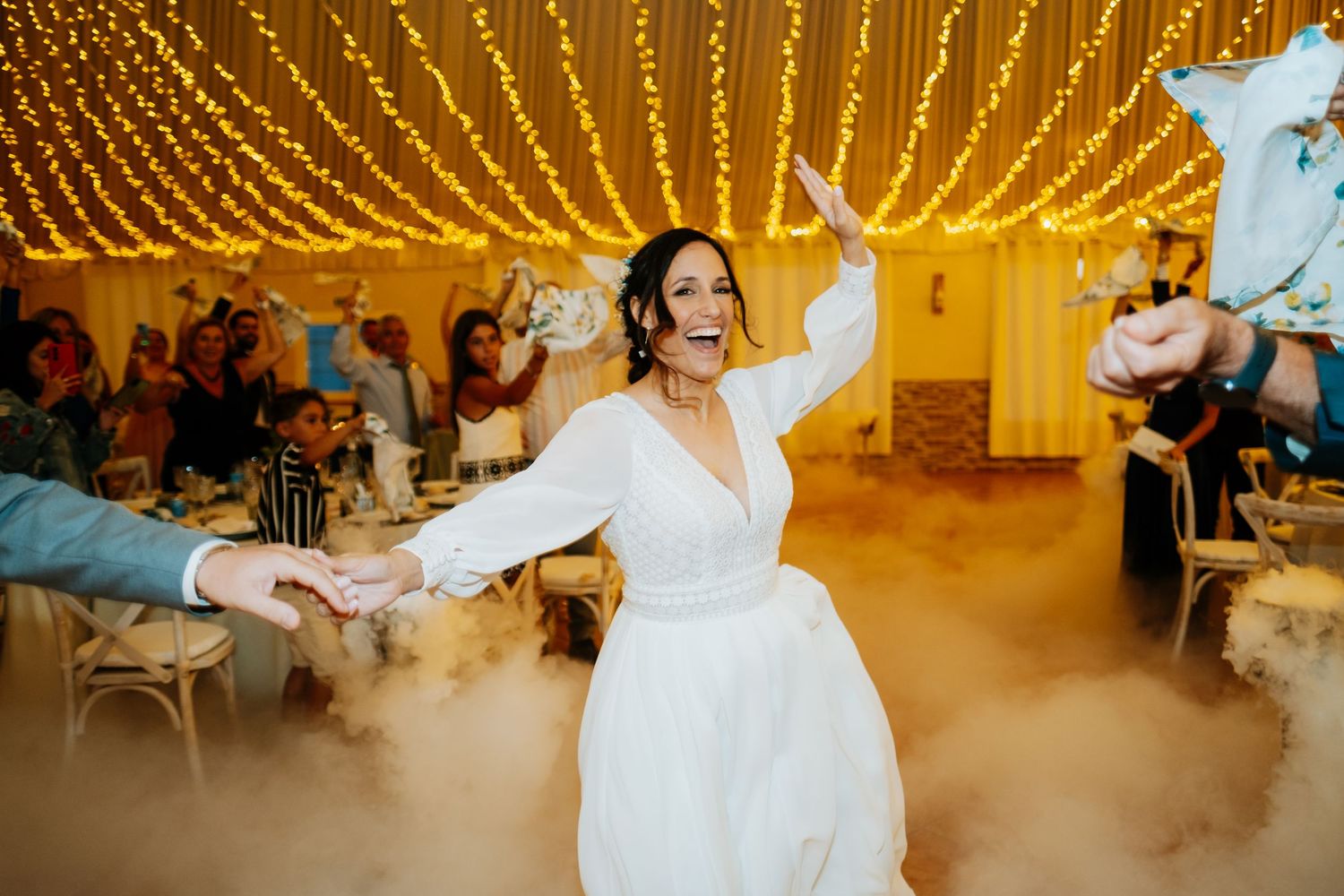 Bride and groom share their first dance surrounded by guests in a warmly lit reception venue with glowing lights overhead.