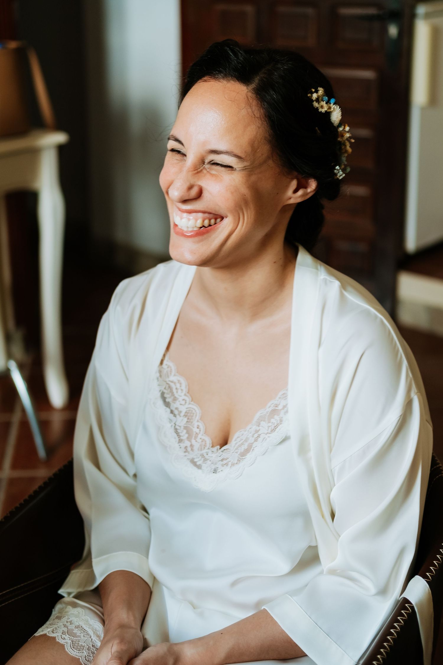 A person in a white robe smiles while having makeup applied during wedding preparations.