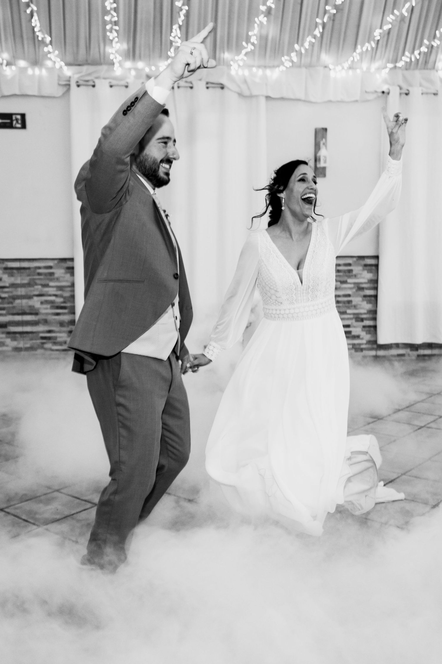 Couple dancing in fog-filled reception venue with string lights overhead in black and white.