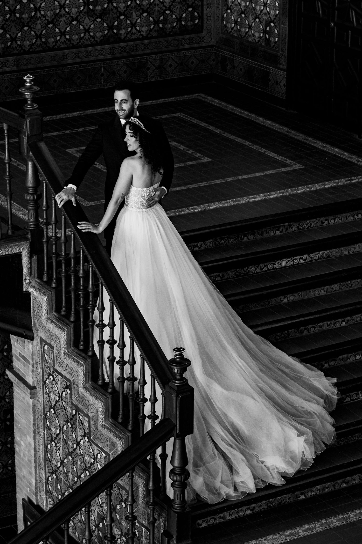 A romantic black and white photo of a couple in wedding attire ascending an ornate vintage staircase with a flowing dress train.