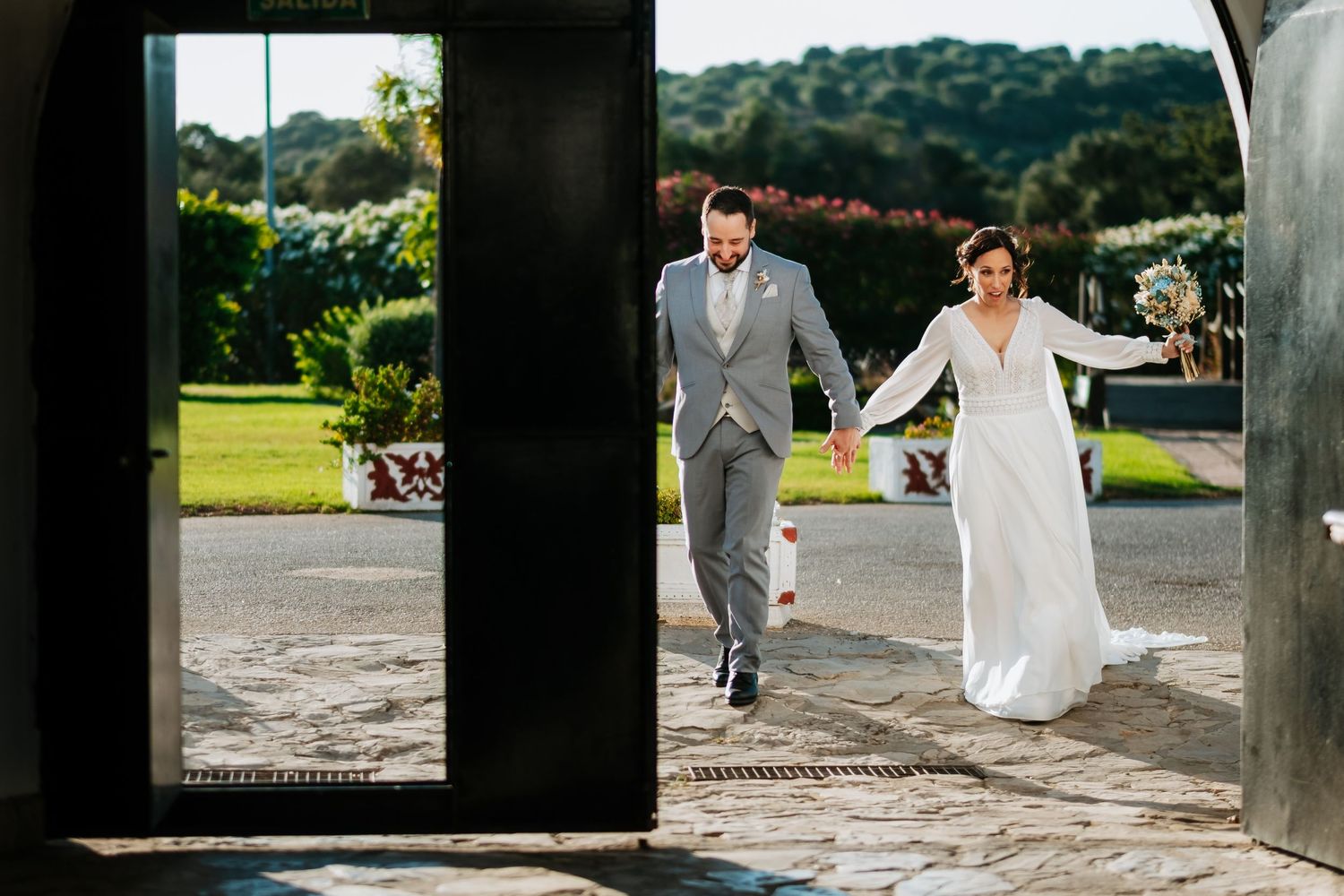 Newlyweds walk hand in hand through a garden pathway surrounded by lush greenery on their wedding day.