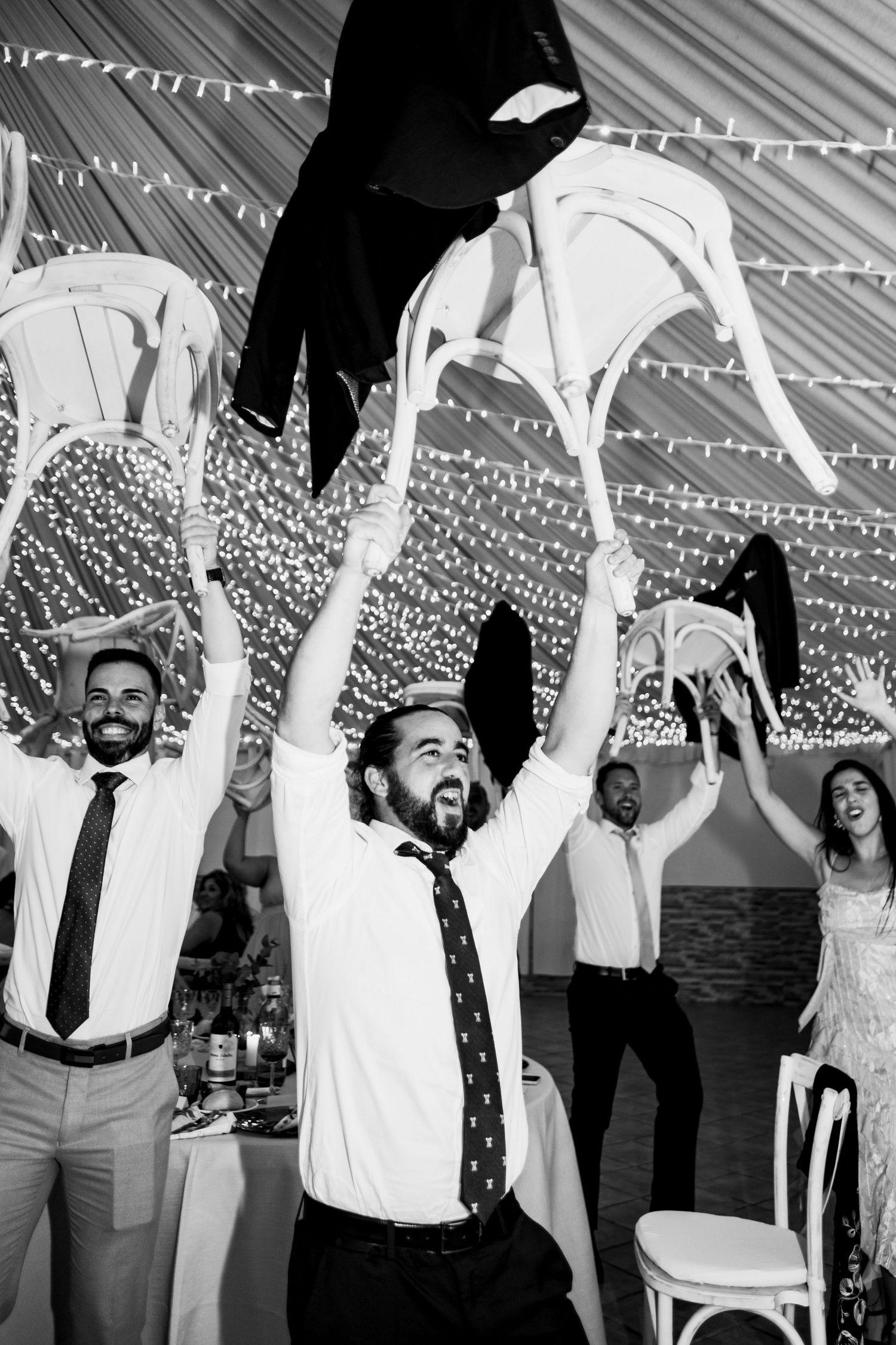 Black and white photo of wedding guests celebrating with raised arms under a tent ceiling.