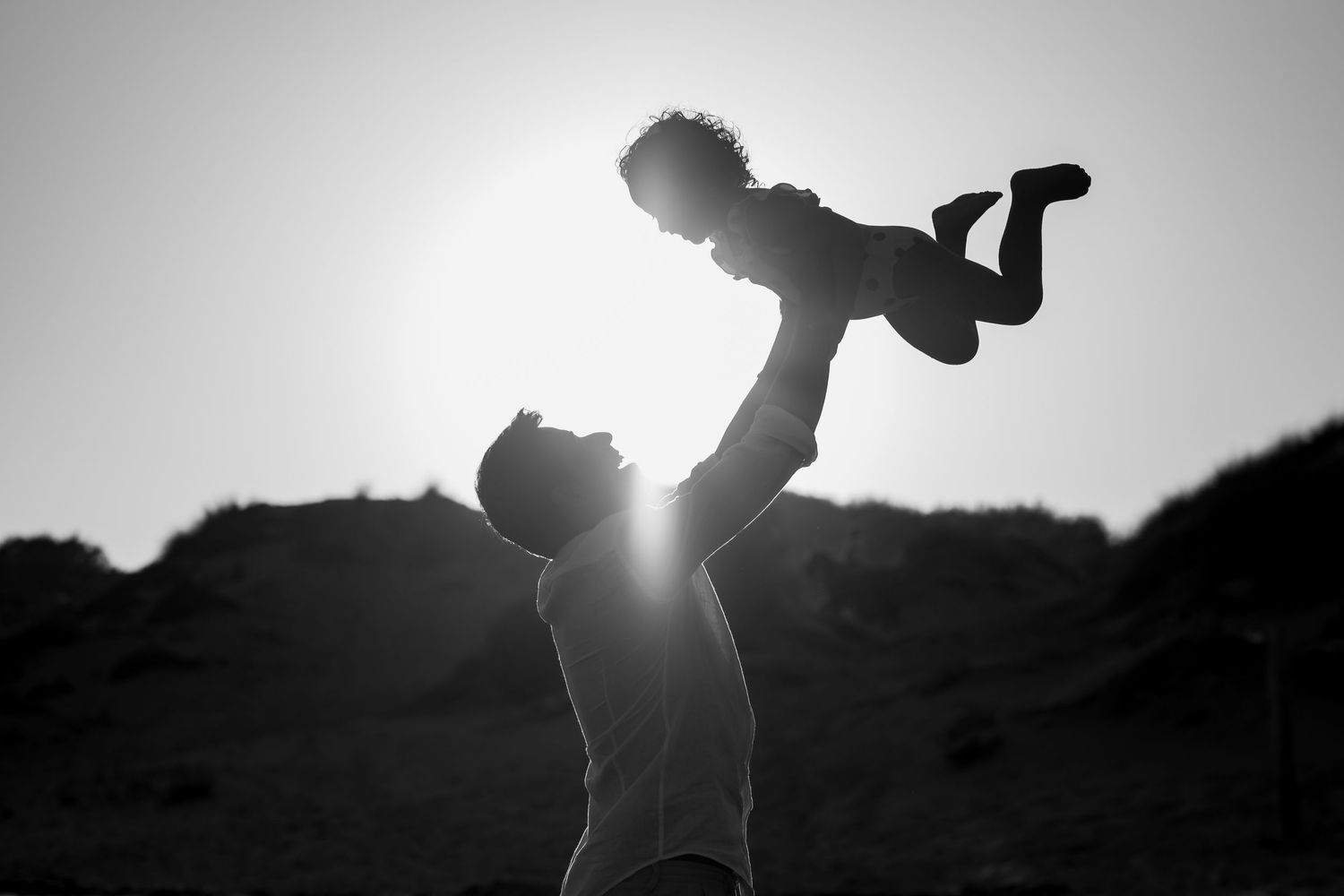 Black and white silhouette of holding a child high in the air during sunset with sun flare peeking through.