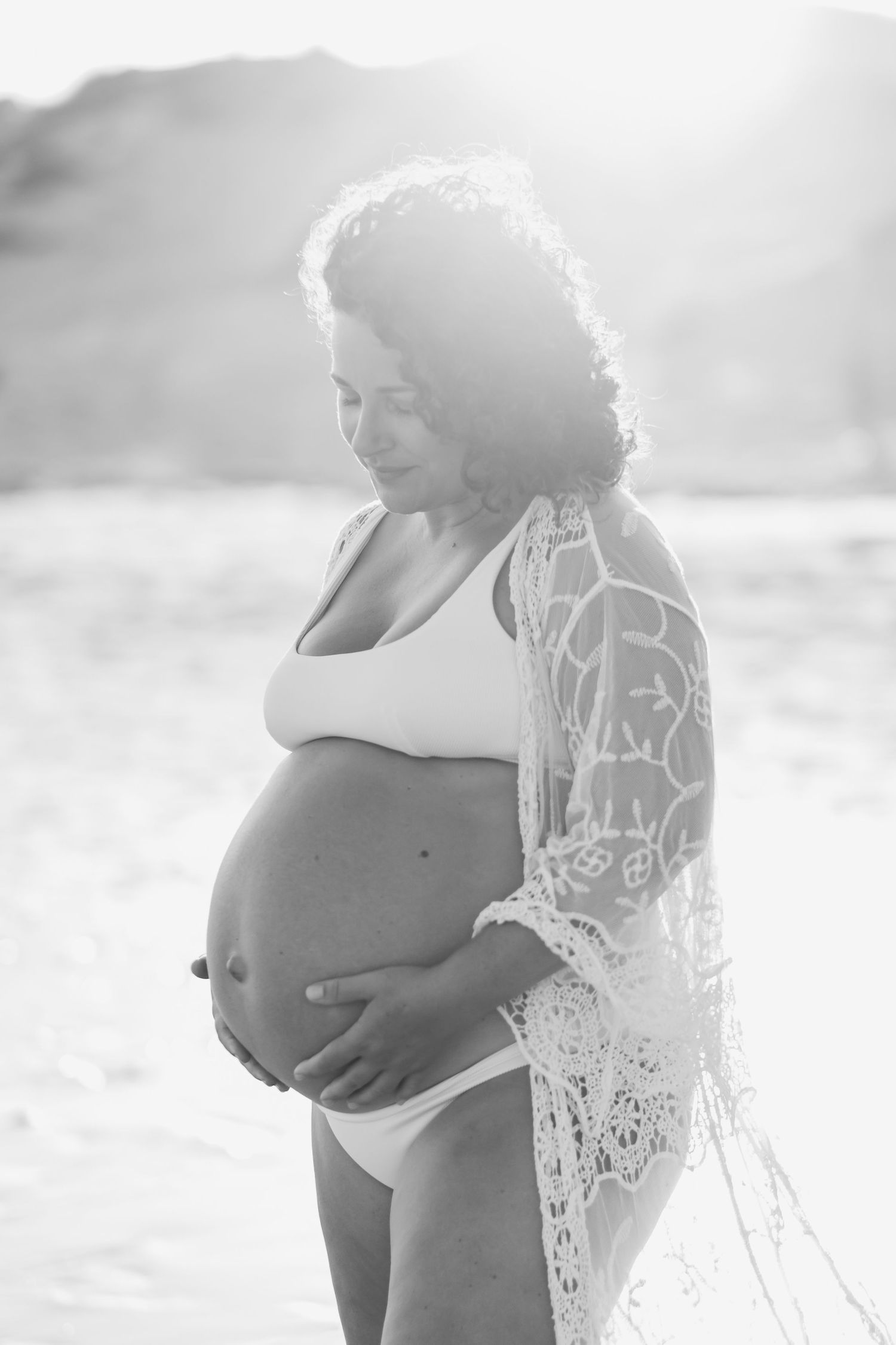 Black and white maternity photo series shows silhouetted figure in lace robe cradling baby bump on beach.