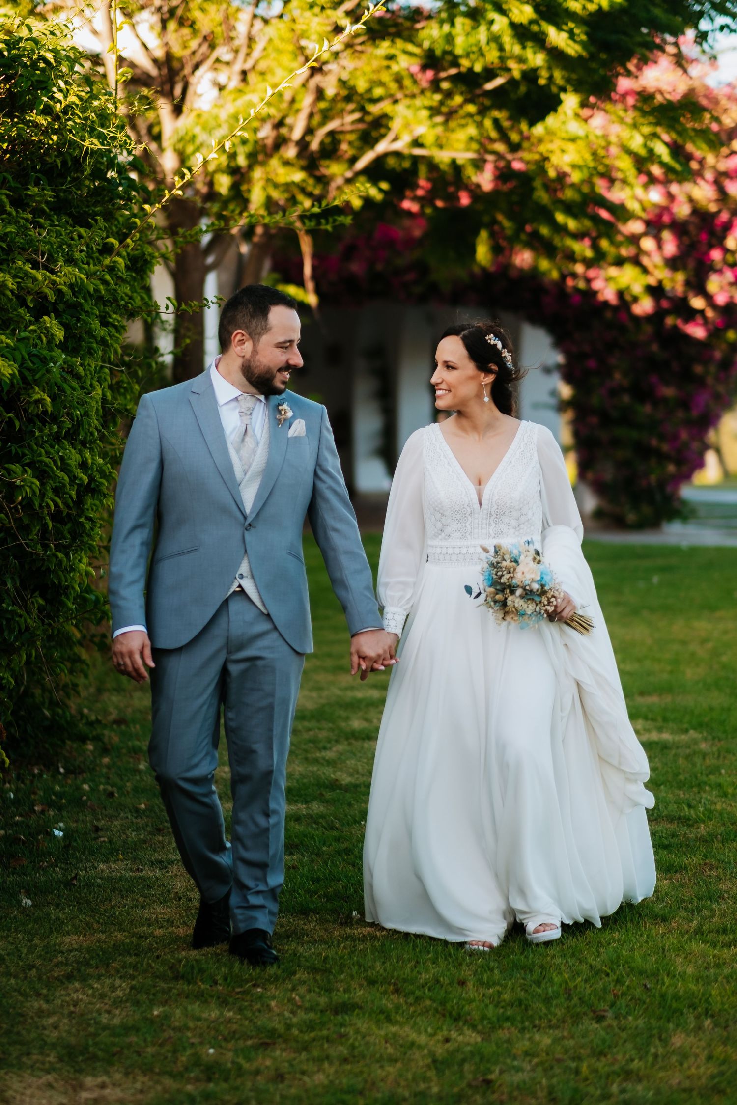 Newlywed couple walking together on green grass with trees and sunlight in romantic outdoor setting.