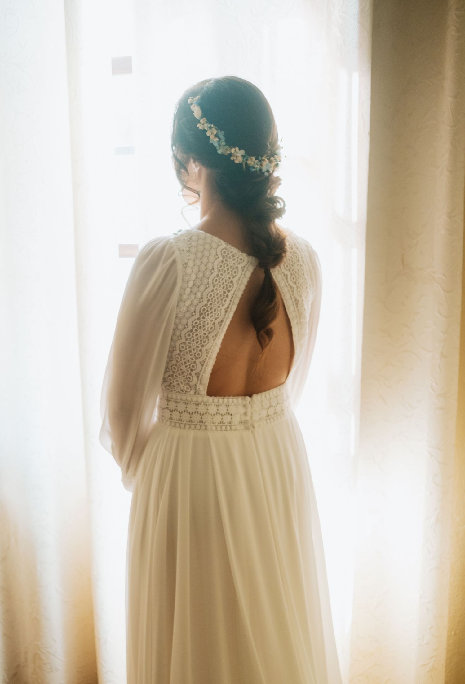 A bride in a flowing white dress with lace details and floral crown stands by a window in soft natural light.
