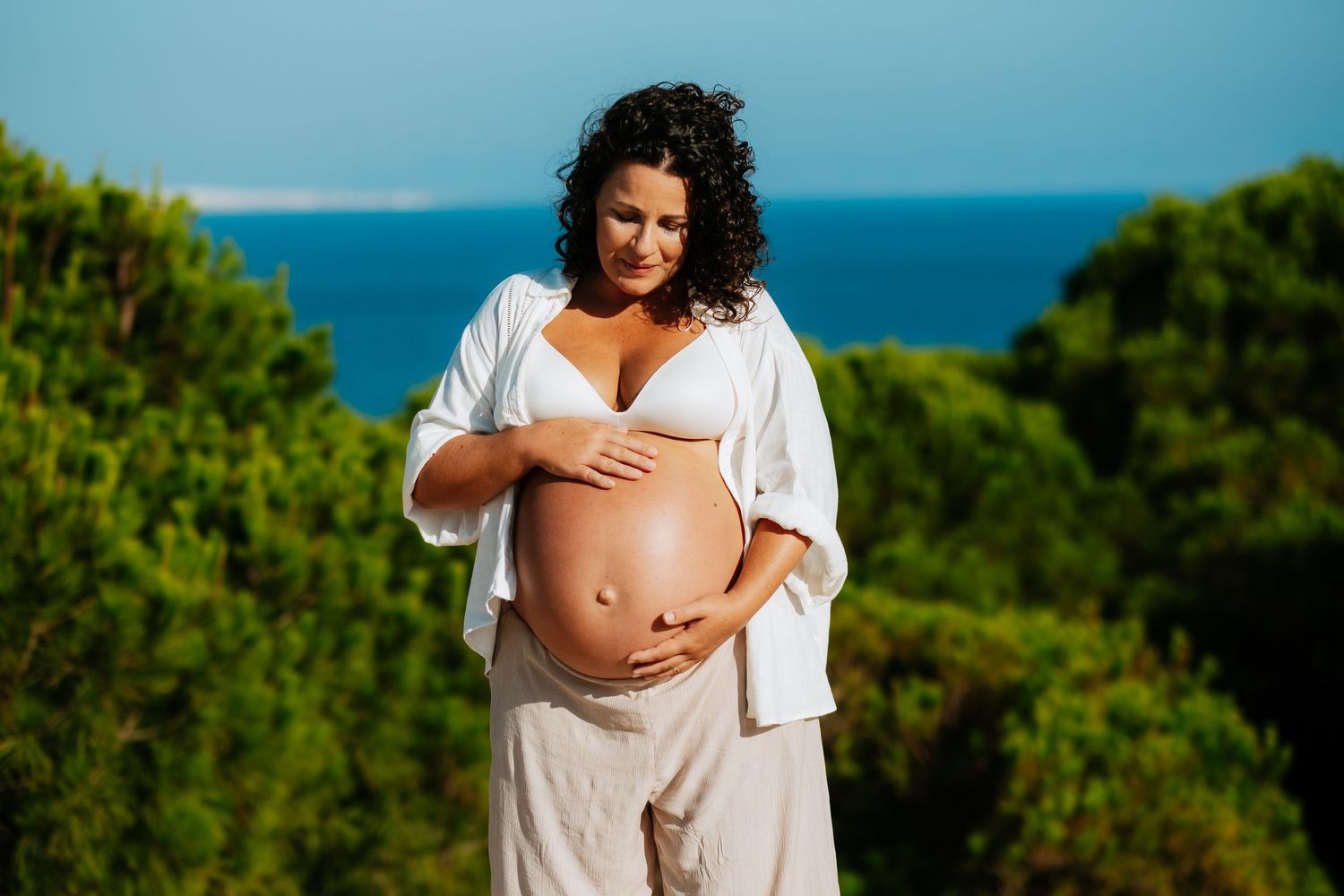Expectant mother in white shirt caresses her baby bump while standing against scenic coastal landscape background.