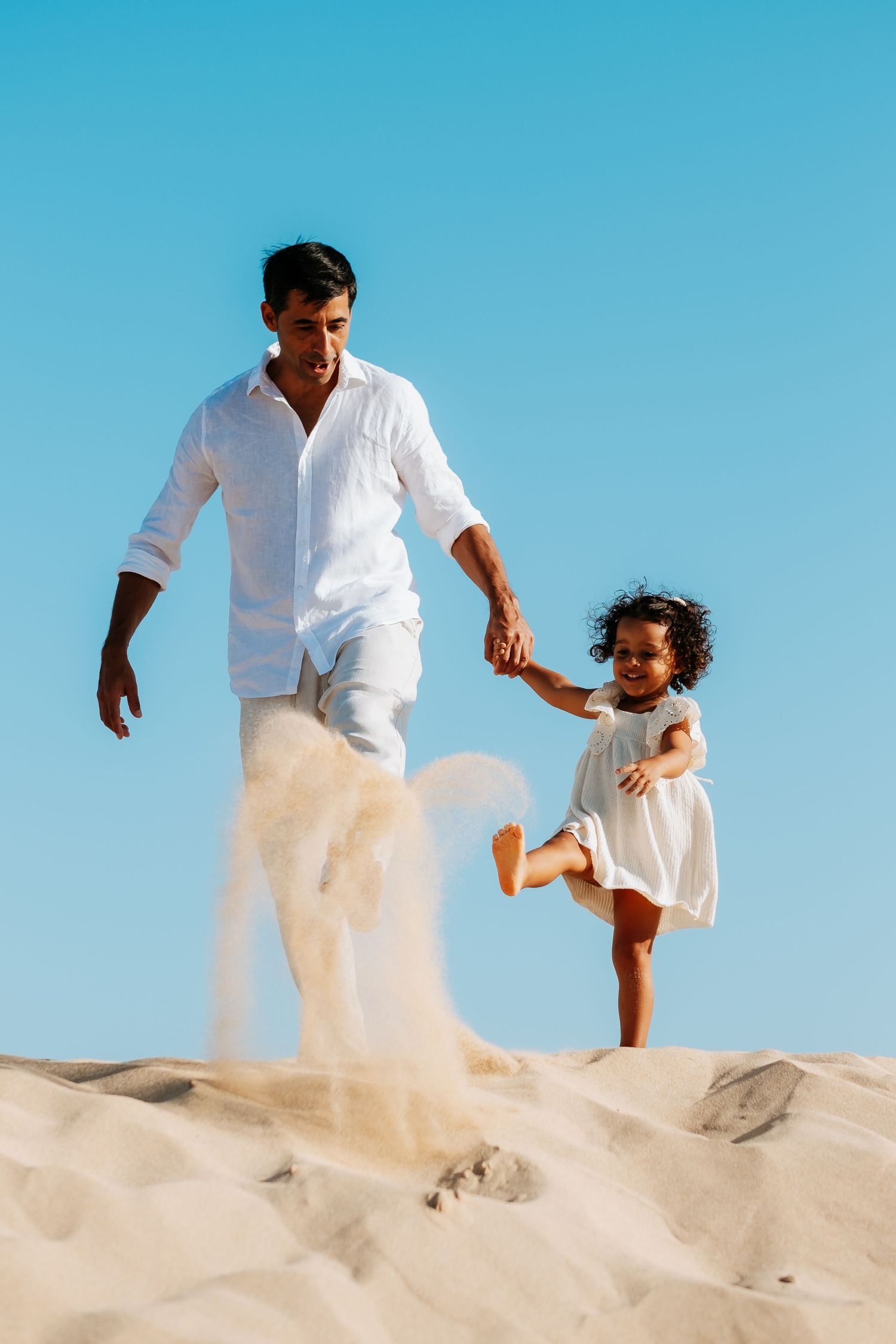 A parent and child in white outfits play together on sandy dunes against a bright blue sky.