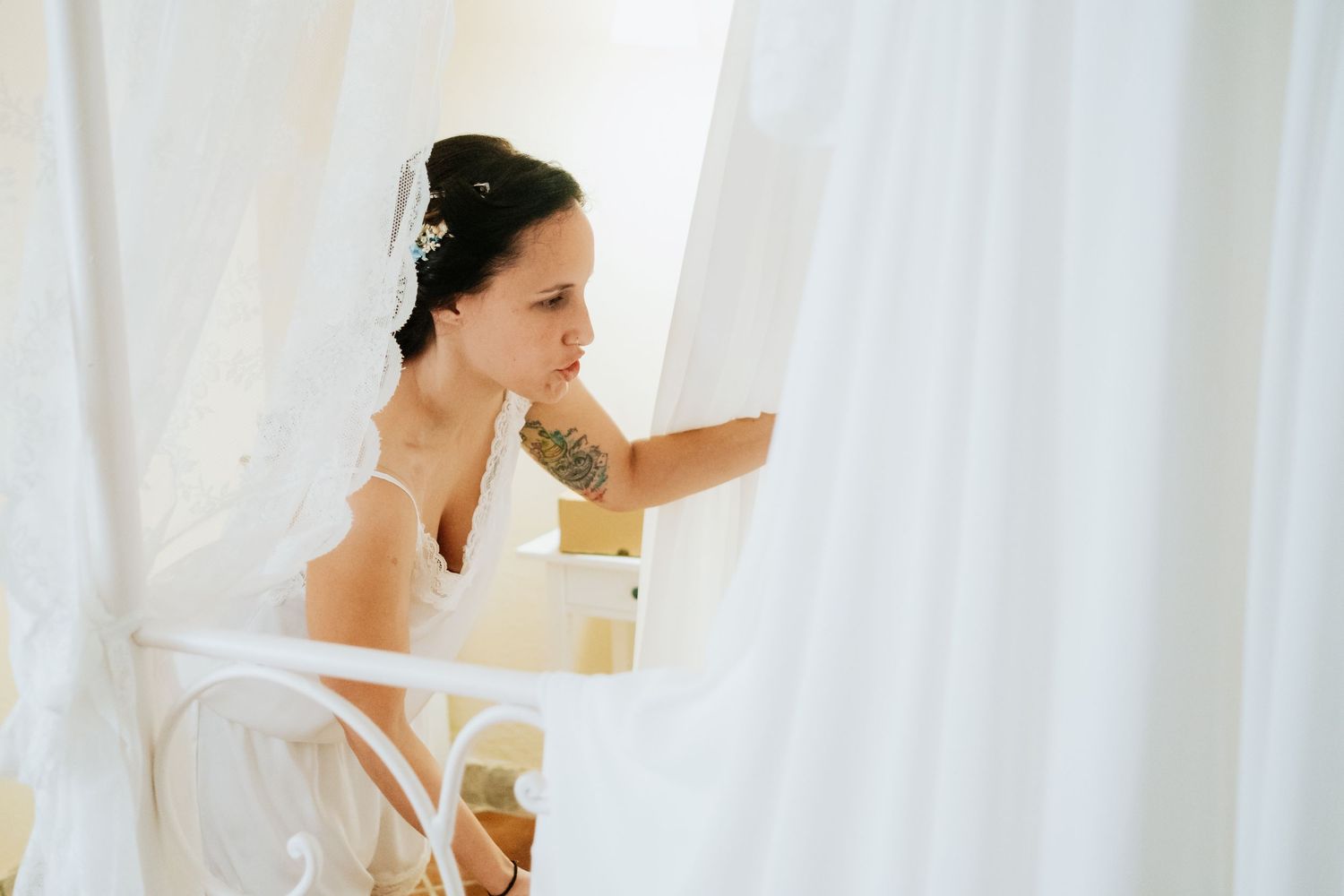 Bride in white getting ready behind sheer curtains in natural window light.