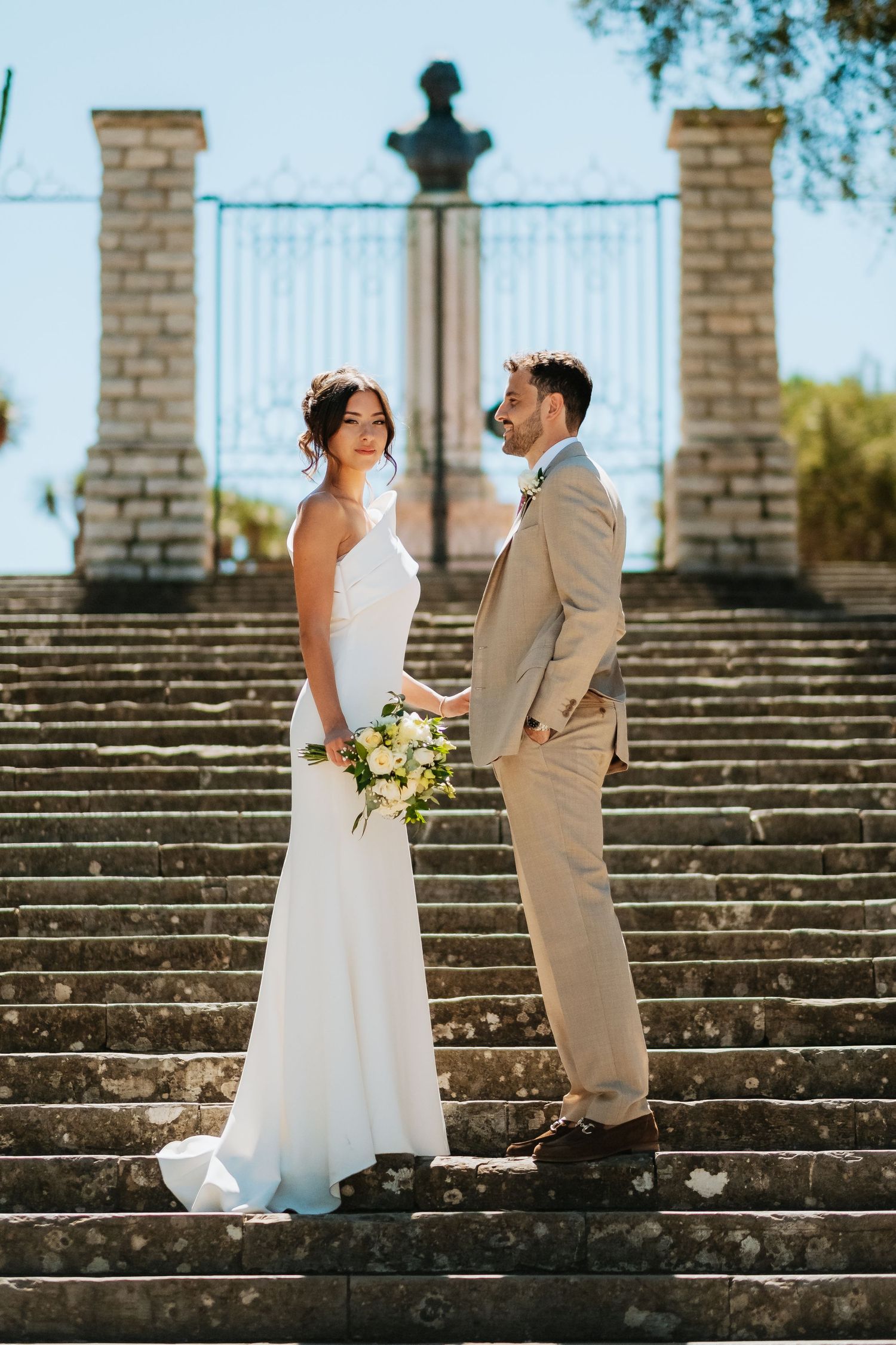 Wedding couple poses on stone steps outdoors in elegant attire on sunny day.