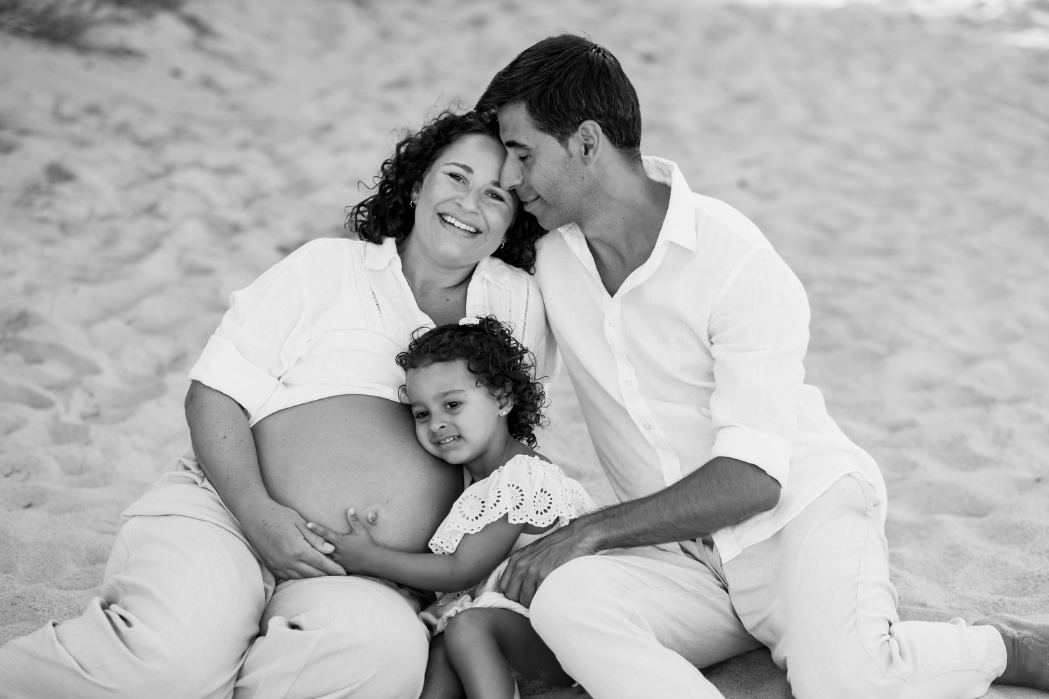 Black and white photo of intimate family moment on beach with expecting parents and child.