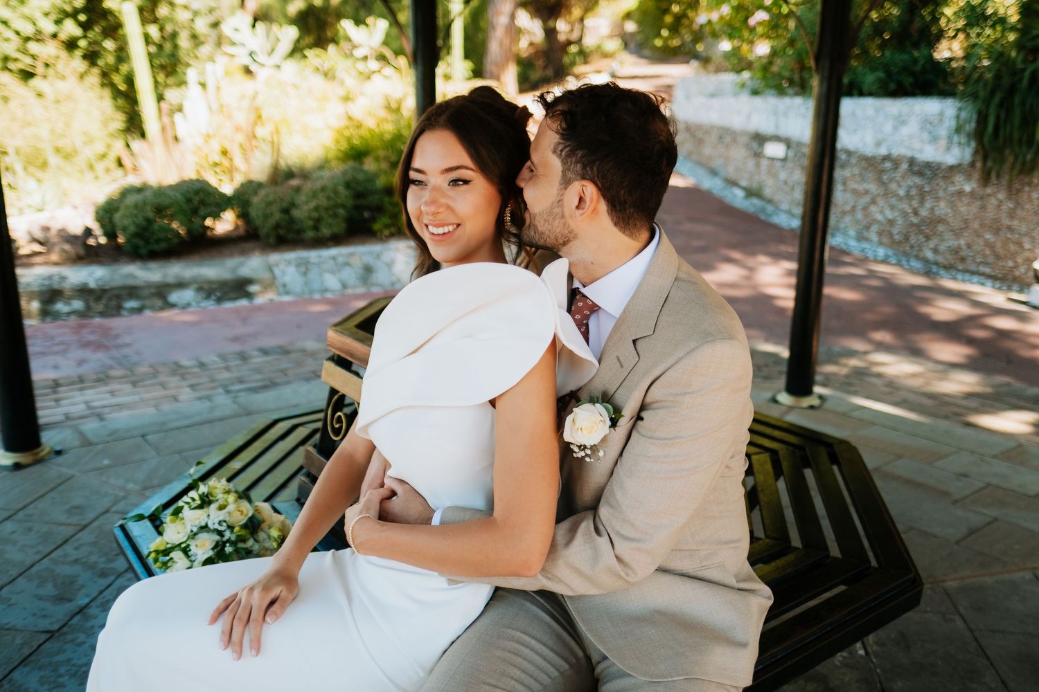 Newlyweds sitting close together on a park bench in dappled sunlight surrounded by greenery.