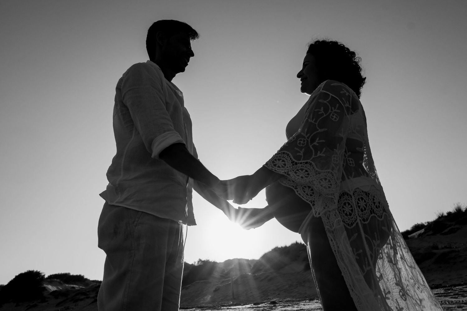 Silhouetted couple holds hands at sunset during a romantic beachside maternity photoshoot.