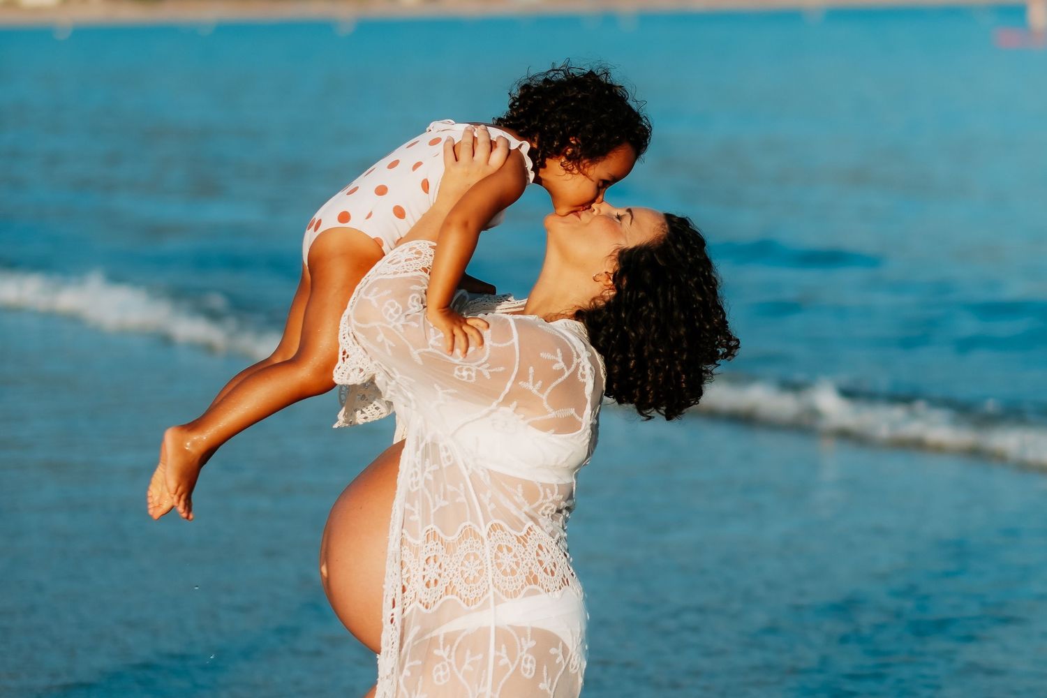 Two people share a tender moment in white lace on a beach at sunset with turquoise ocean waves in the background.