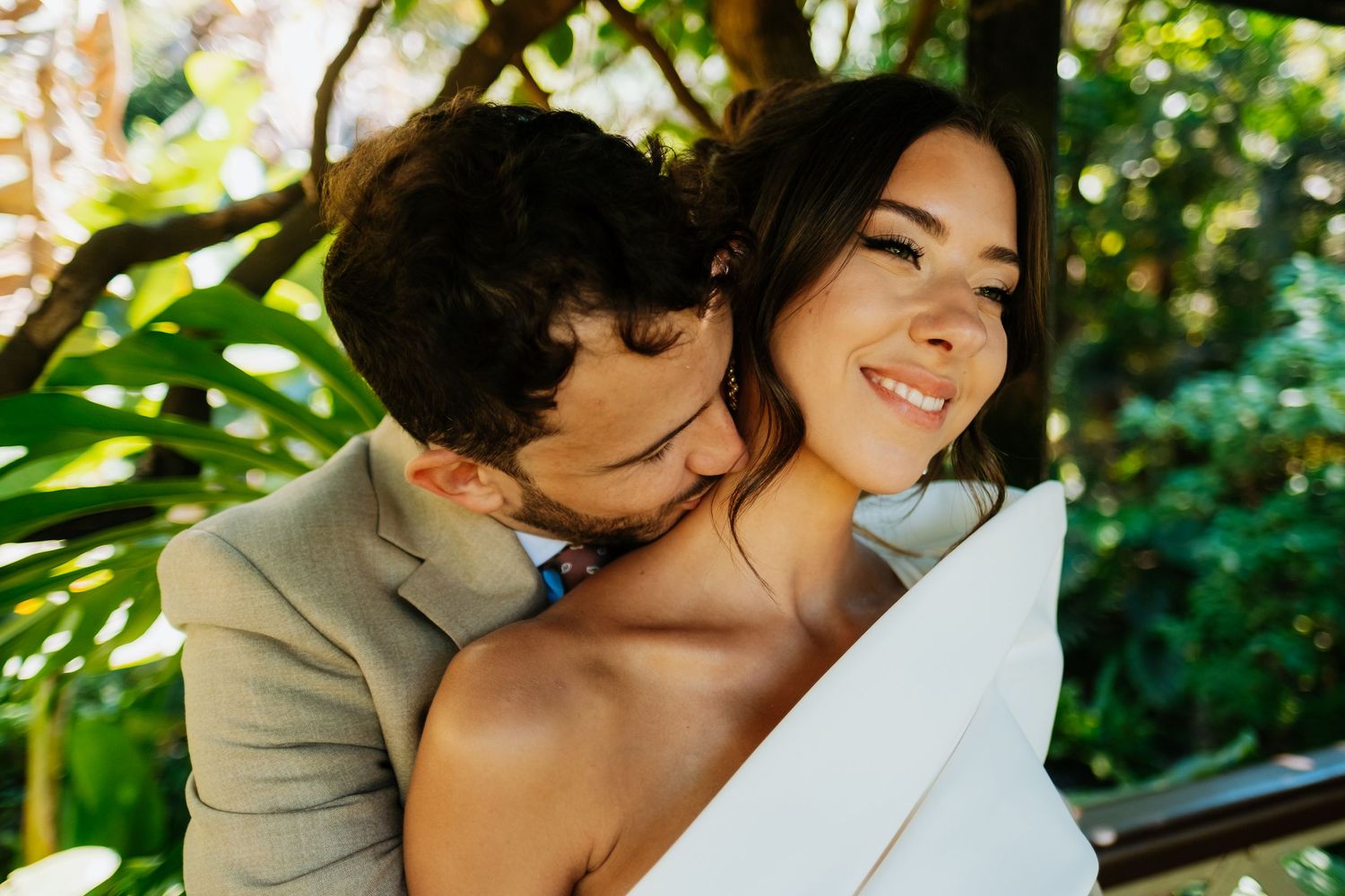 Newlyweds embracing in lush garden setting with natural sunlight filtering through trees.