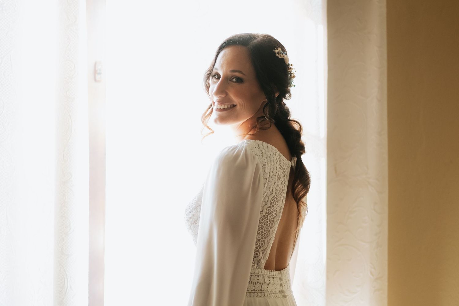 A bride in a white wedding dress stands by a window in soft natural light during a wedding preparation moment.