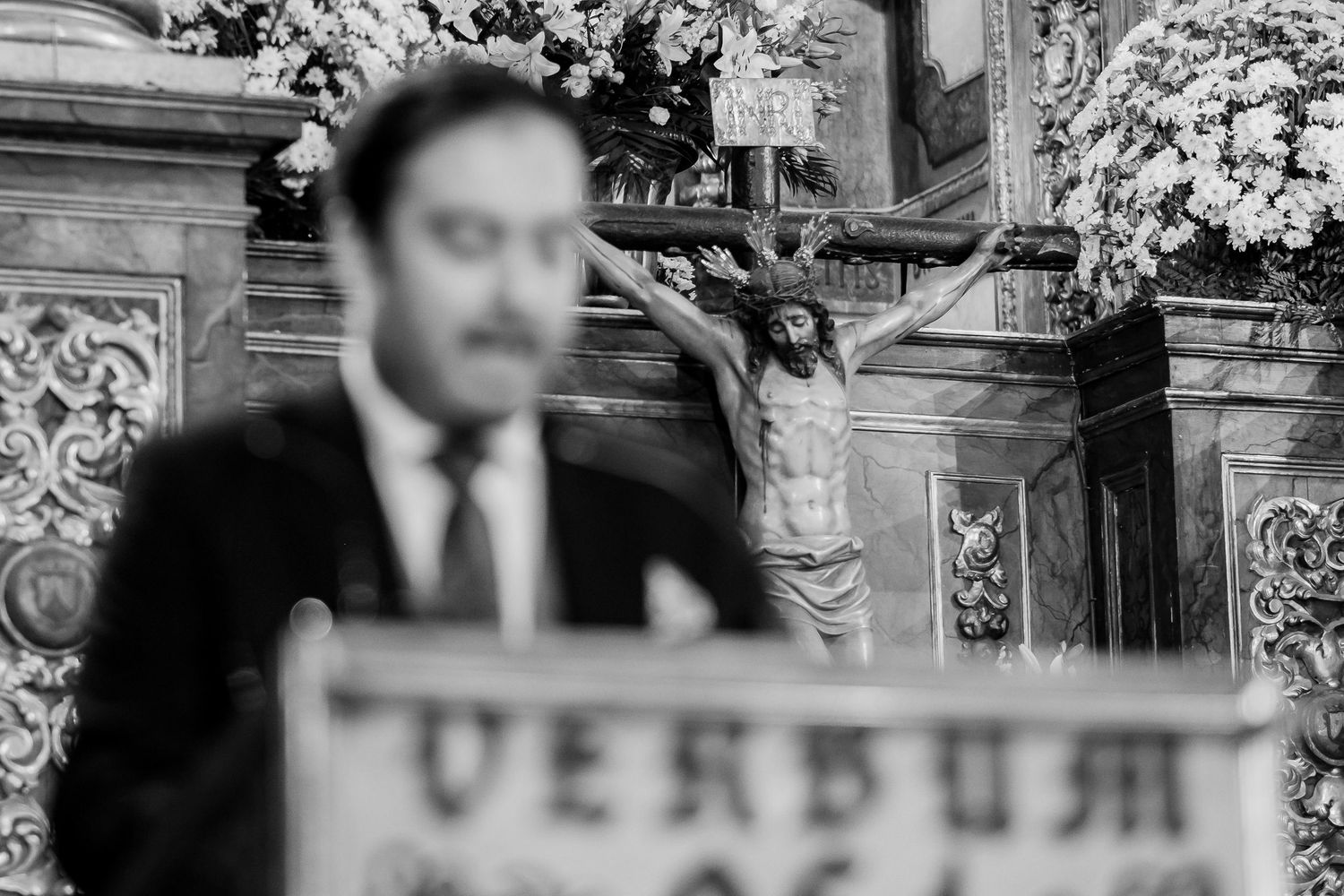 Fotografía de boda en blanco y negro en la Basílica del Carmen Jerez, desenfoque selectivo del lector en el ambón y enfoque en el crucifijo