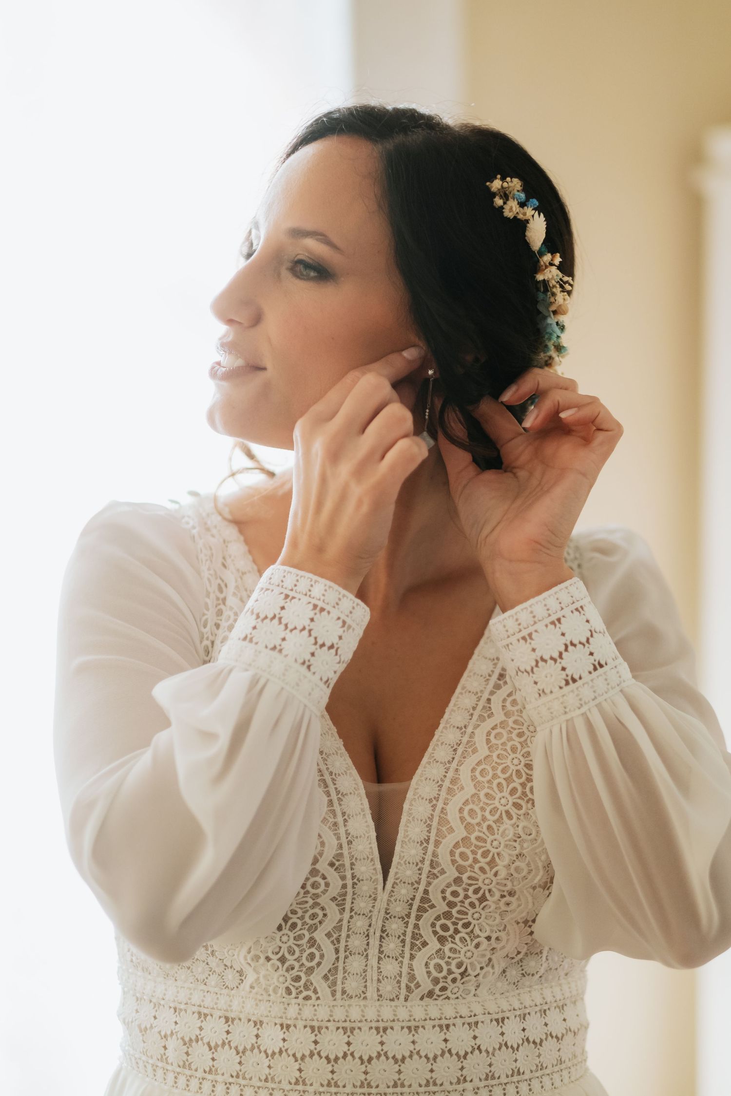 Bride in white lace dress getting ready while adjusting earrings in morning light.