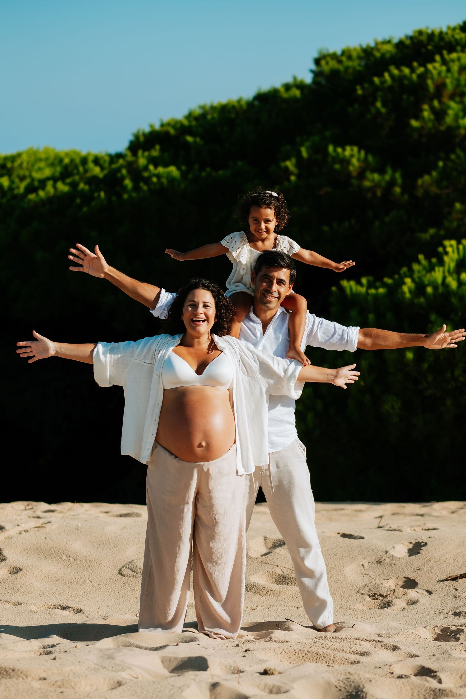Family poses joyfully on beach with arms outstretched while wearing white outfits against green foliage.