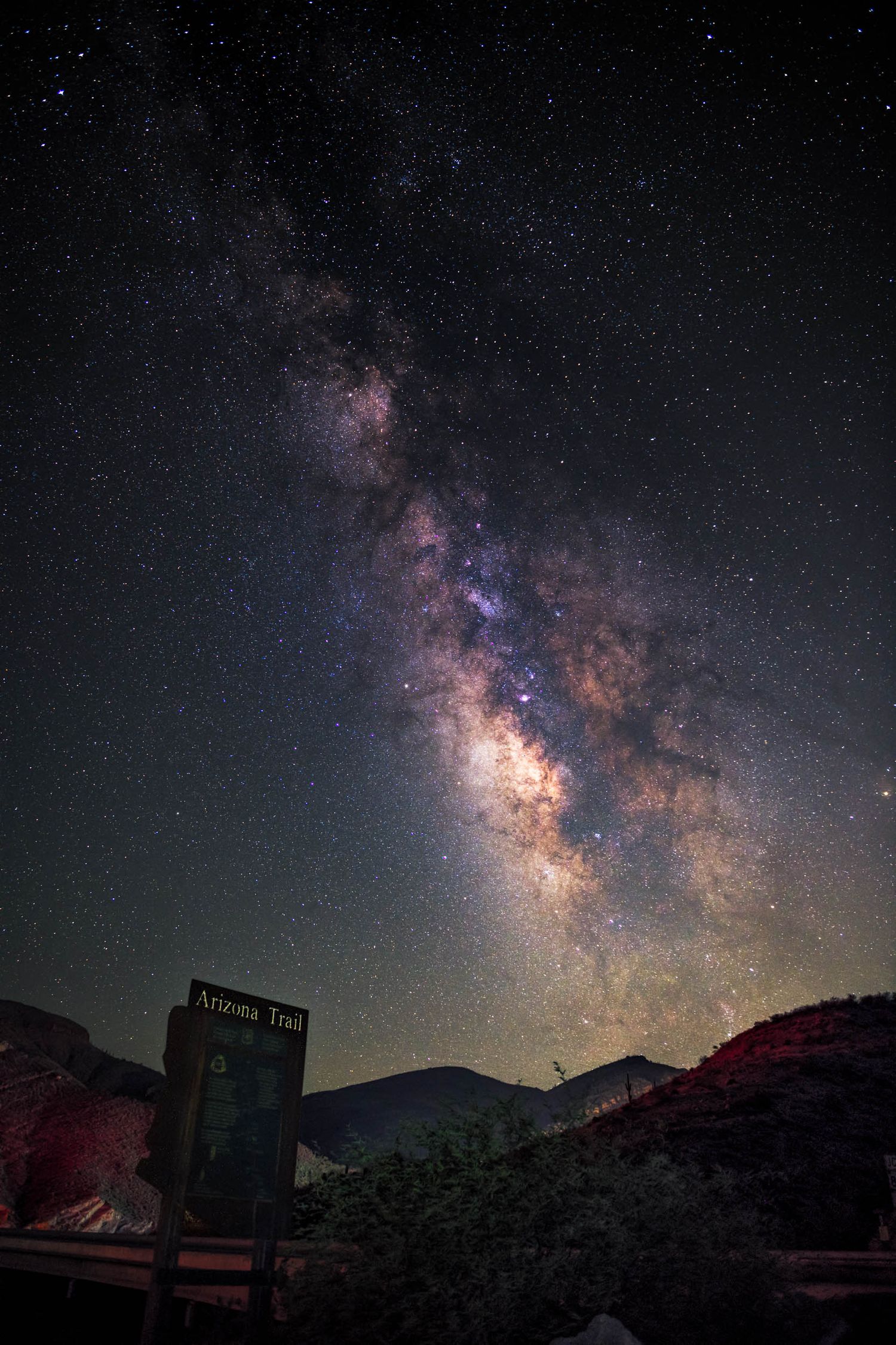 A Milky Way photograph above an Arizona Trail sign and mountain horizon, combining night sky, adventure, and desert landscape.