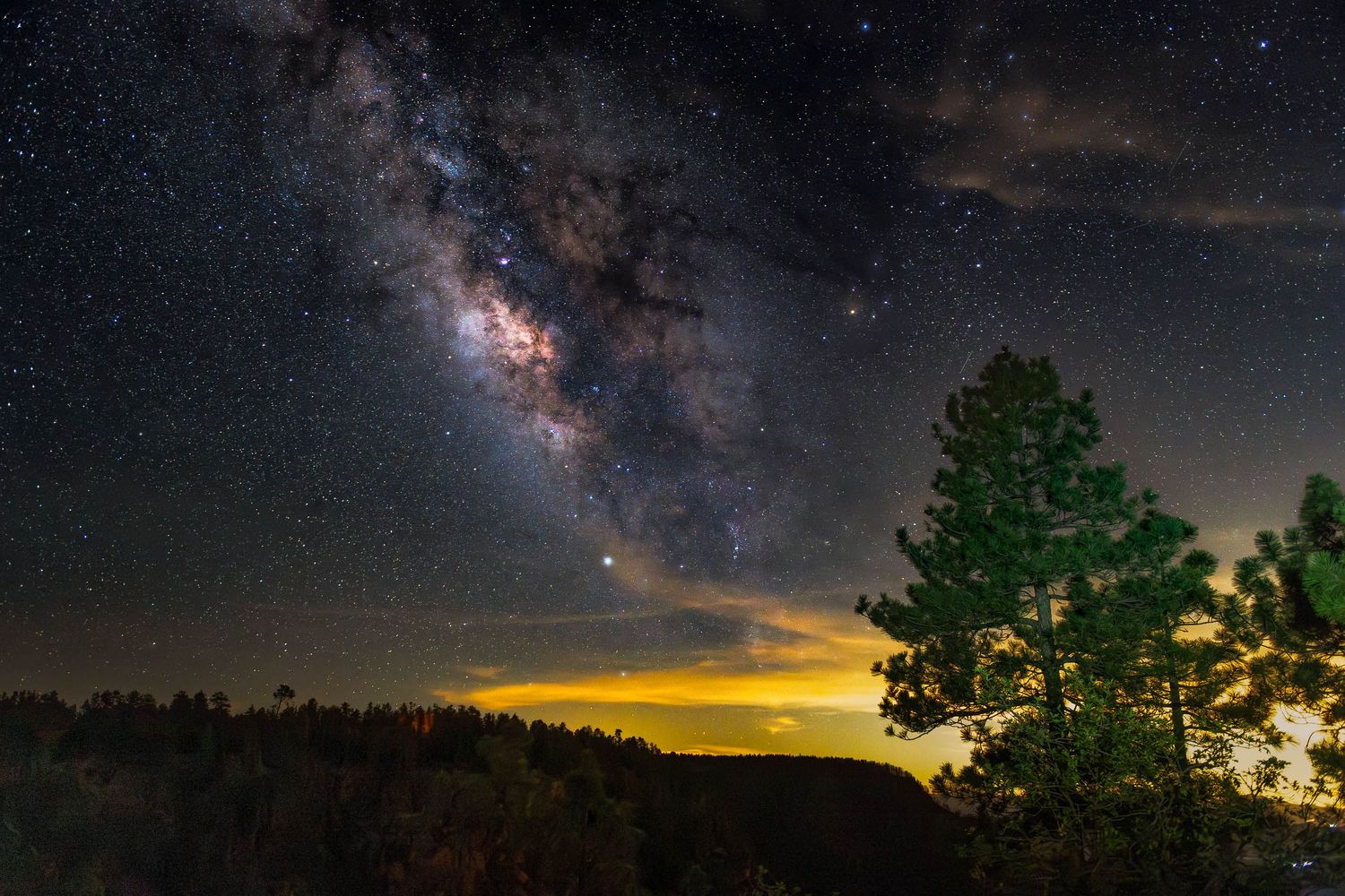 Mogollon Rim Milky Way nightscape above pine silhouettes with soft horizon glow.