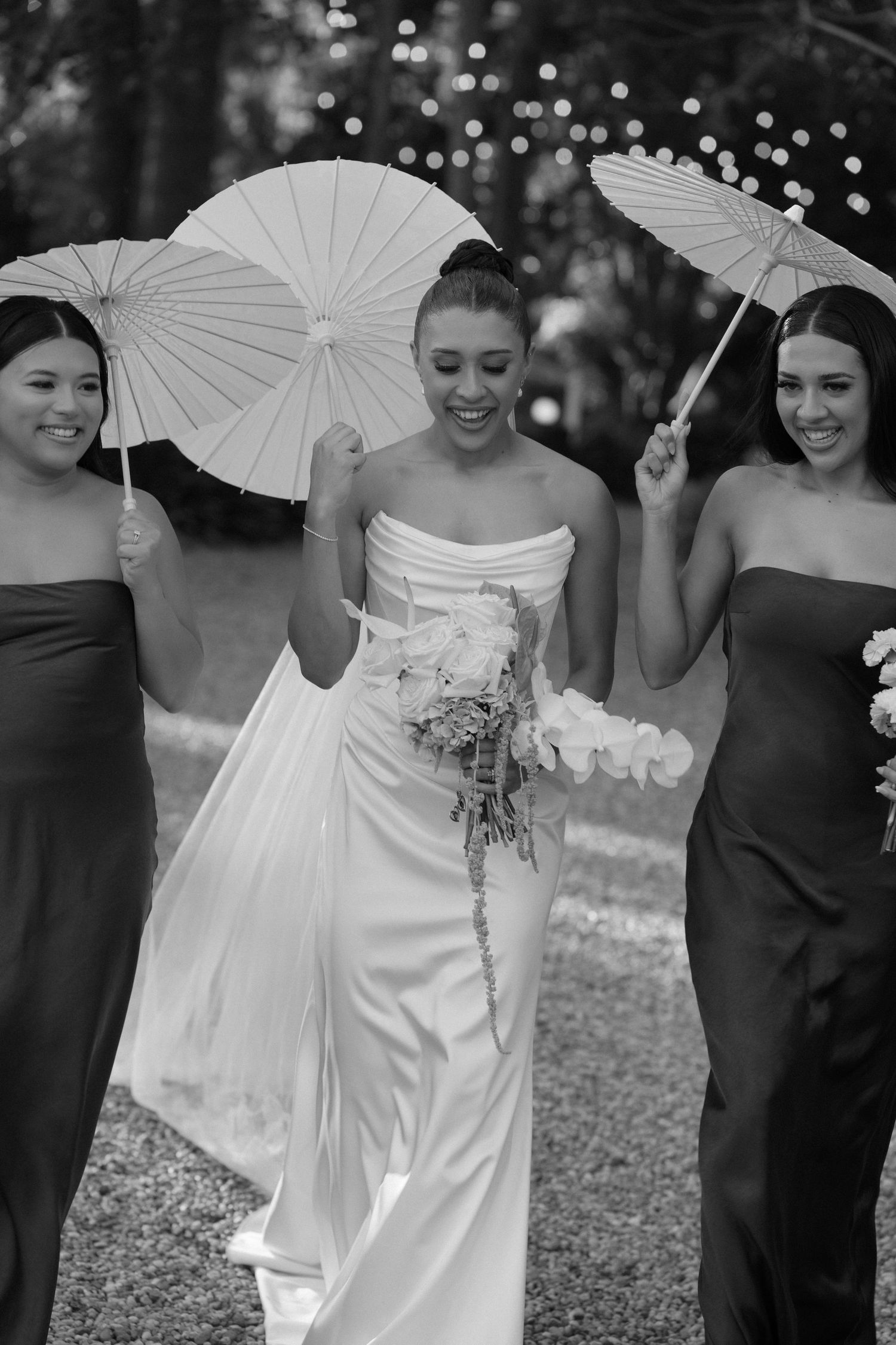 Black and white wedding photo showing bride in sleek white gown with bridesmaids holding paper parasols outdoors.