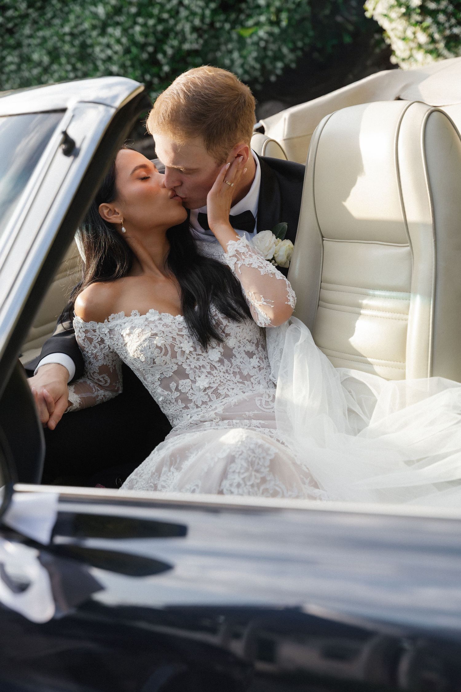 Newlyweds share a romantic kiss in the backseat of a classic car, with bride wearing an elegant off-shoulder lace gown.