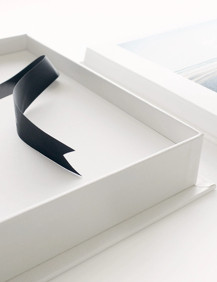 Open white rigid gift box with a black satin ribbon draped inside, beside a framed print on a bright white surface.