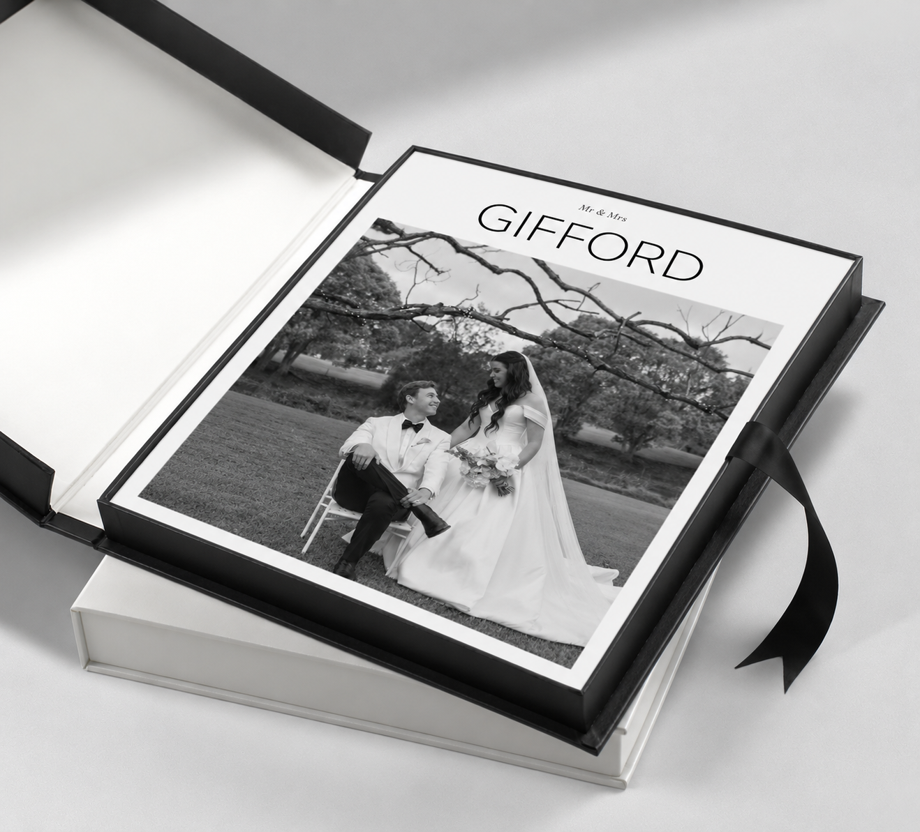 Black and white wedding album titled Gifford open in a black presentation box with ribbon, showing a bride and groom seated outdoors.
