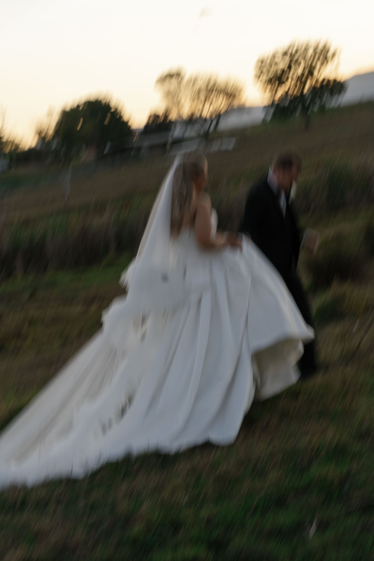 Blurry motion shot of bride in white gown and groom in black suit walking through a field at dusk.