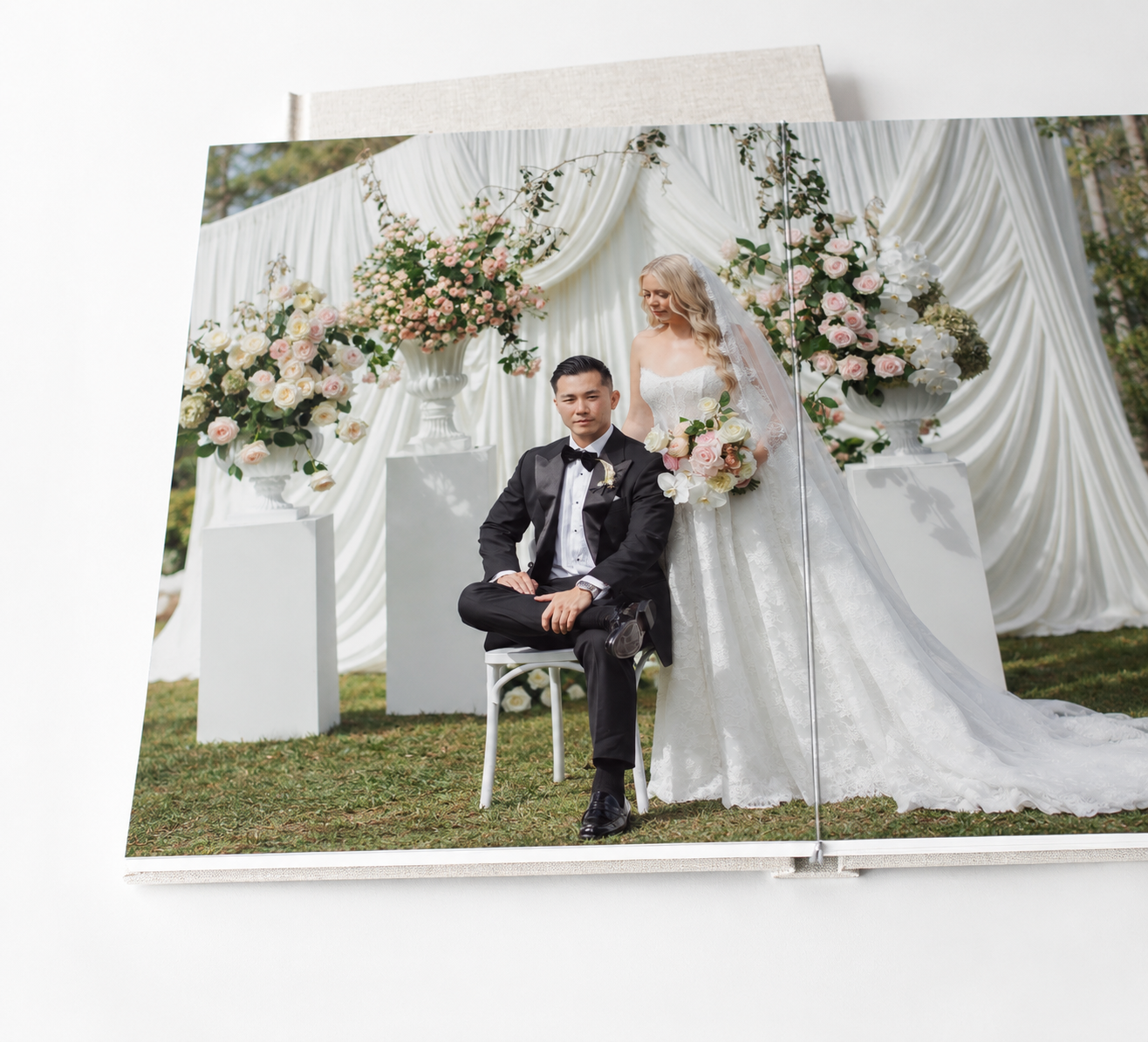 Open wedding photo album showing a bride in a white gown holding a bouquet beside a seated groom in a black tuxedo at a floral ceremony.