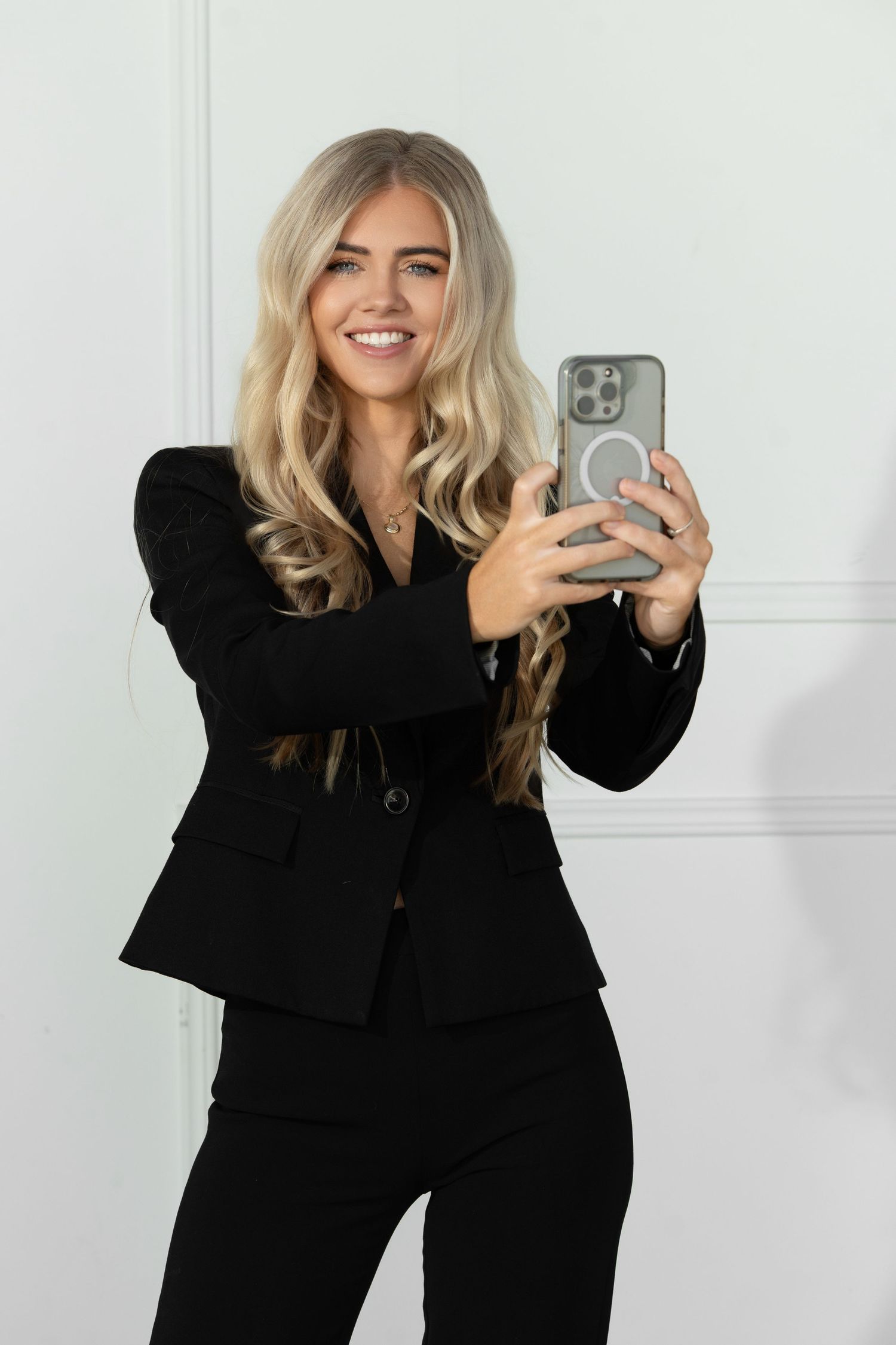 A person wearing a black blazer poses for mirror selfies in four different poses against a white wall.