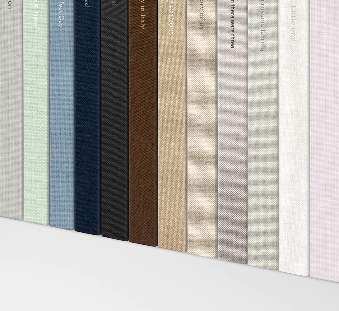 Row of linen-bound book spines in sage green, slate blue, navy, black, brown, tan, gray, and cream with embossed titles on a white shelf.