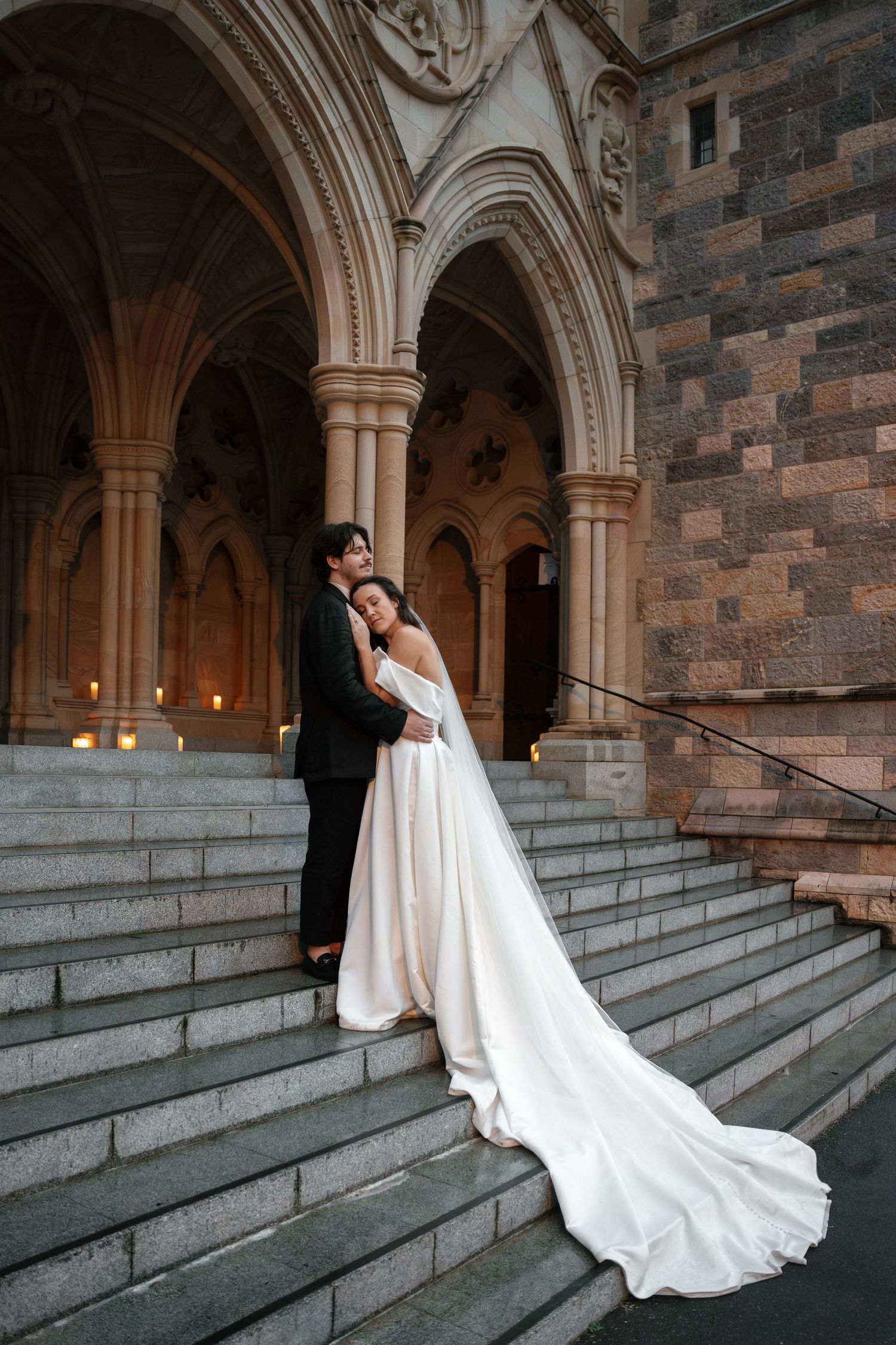 A wedding couple embracing on stone church steps with gothic architecture in the background.