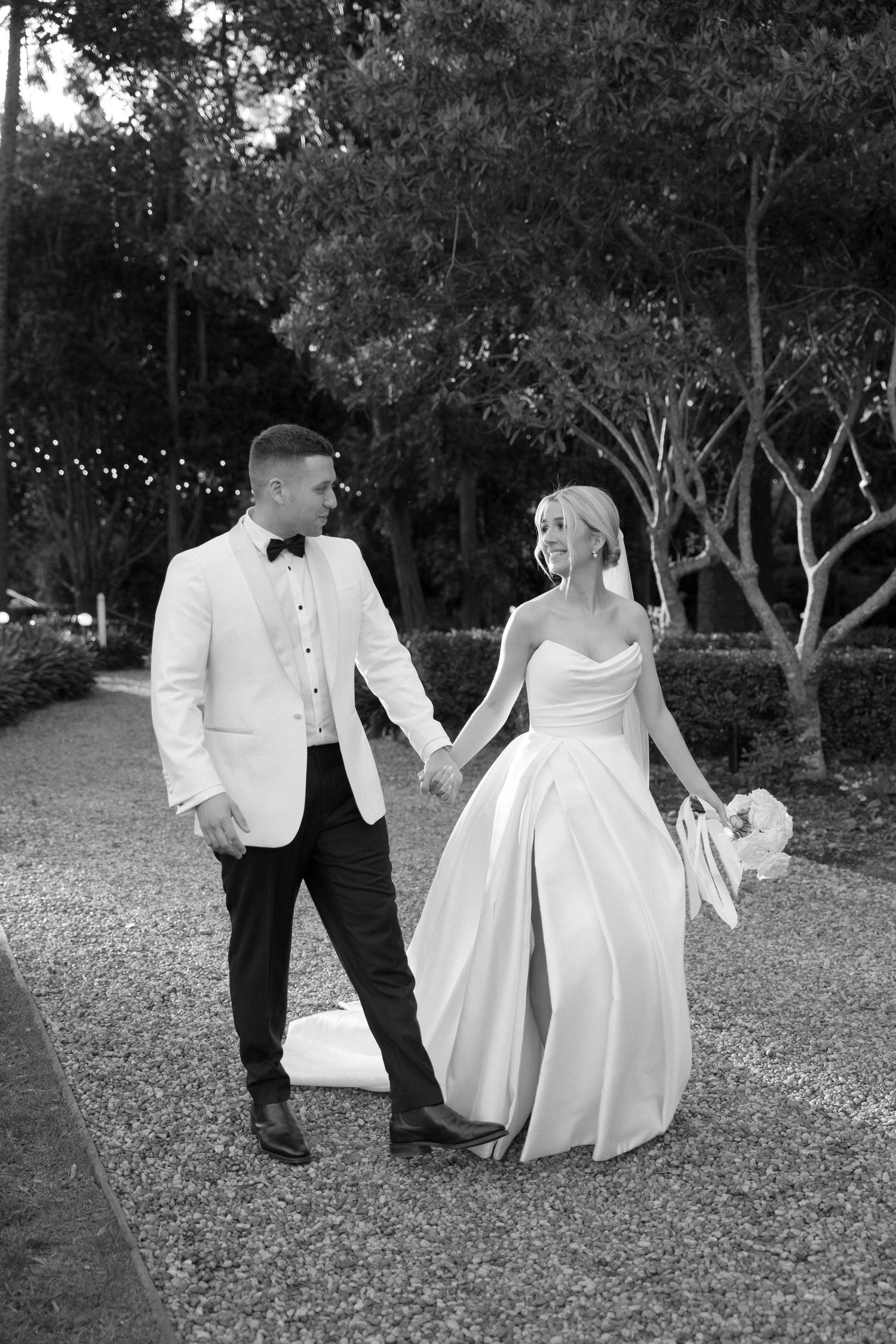 A romantic black and white photo of newlyweds walking together through a garden setting.