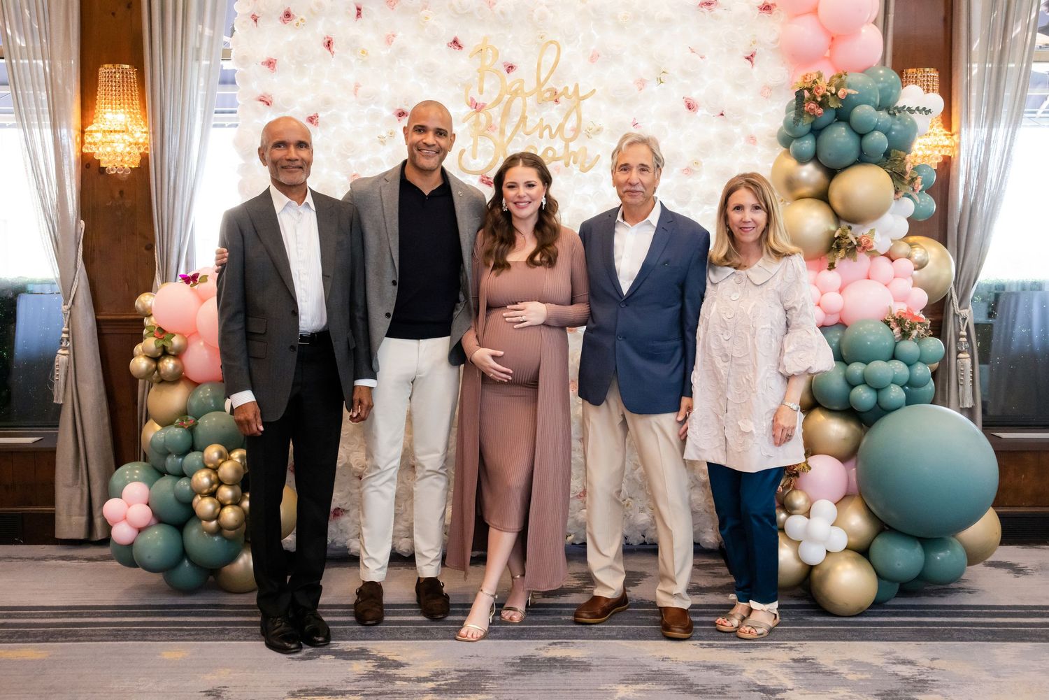 Five guests pose at a baby shower with pink and green balloon arch decorations and a floral backdrop.