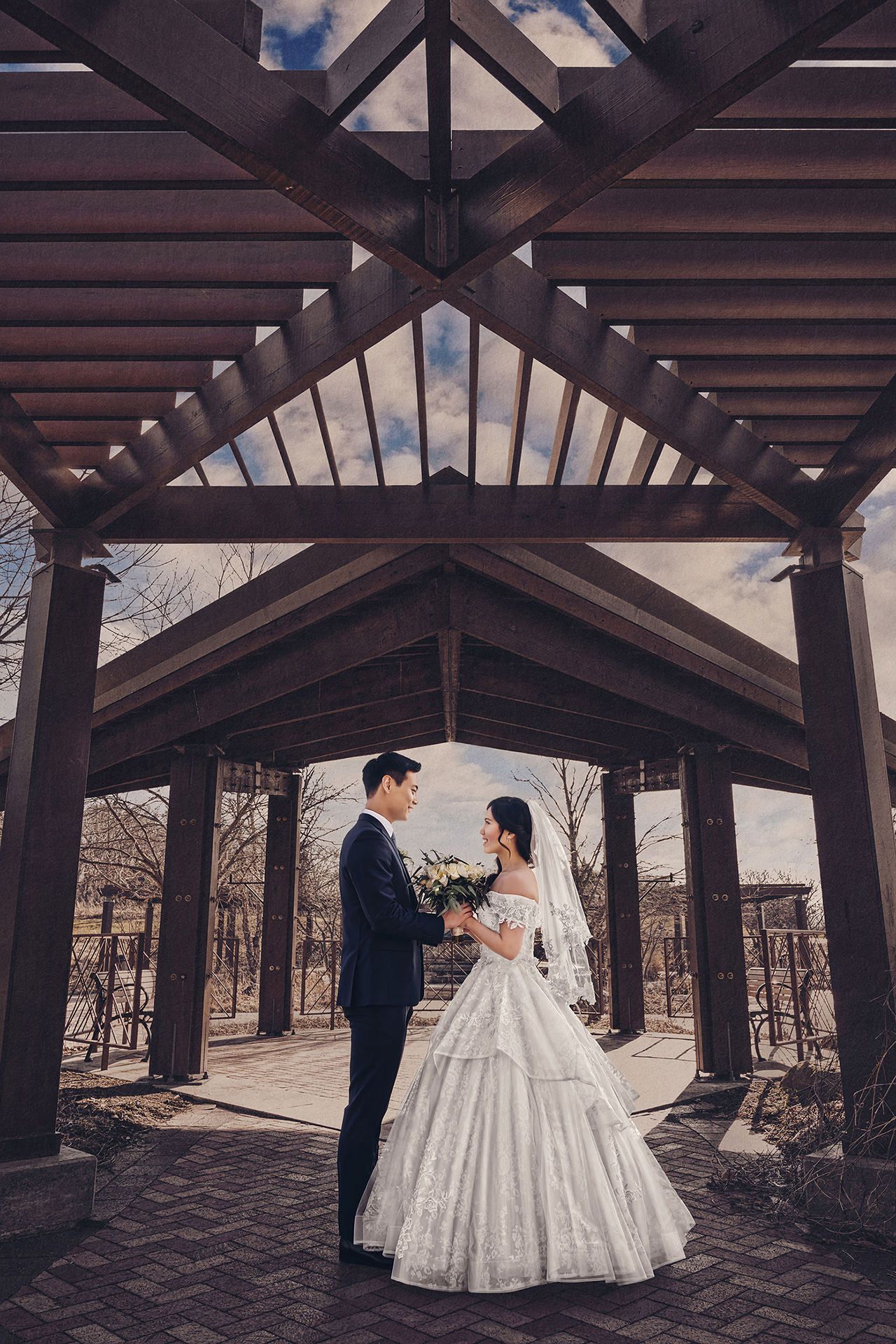 Bride and groom stand facing each other under a wooden pergola, bride in a white ball gown holding a bouquet.