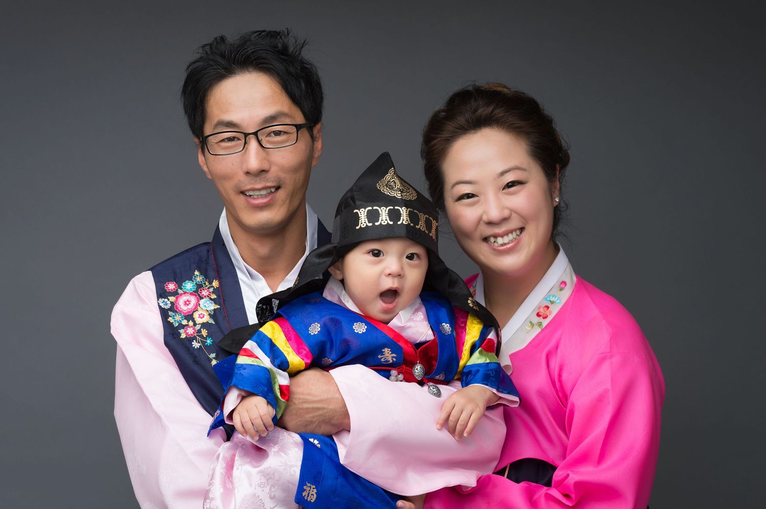 Family of three in traditional Korean hanbok attire, parents holding baby in colorful costume, studio portrait.