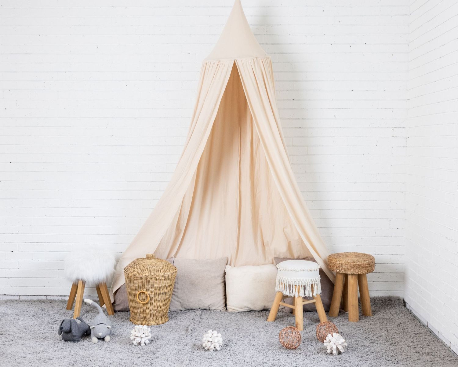 A cozy reading nook with cream canopy tent, wooden stools, woven basket, and plush cushions against a white wall.