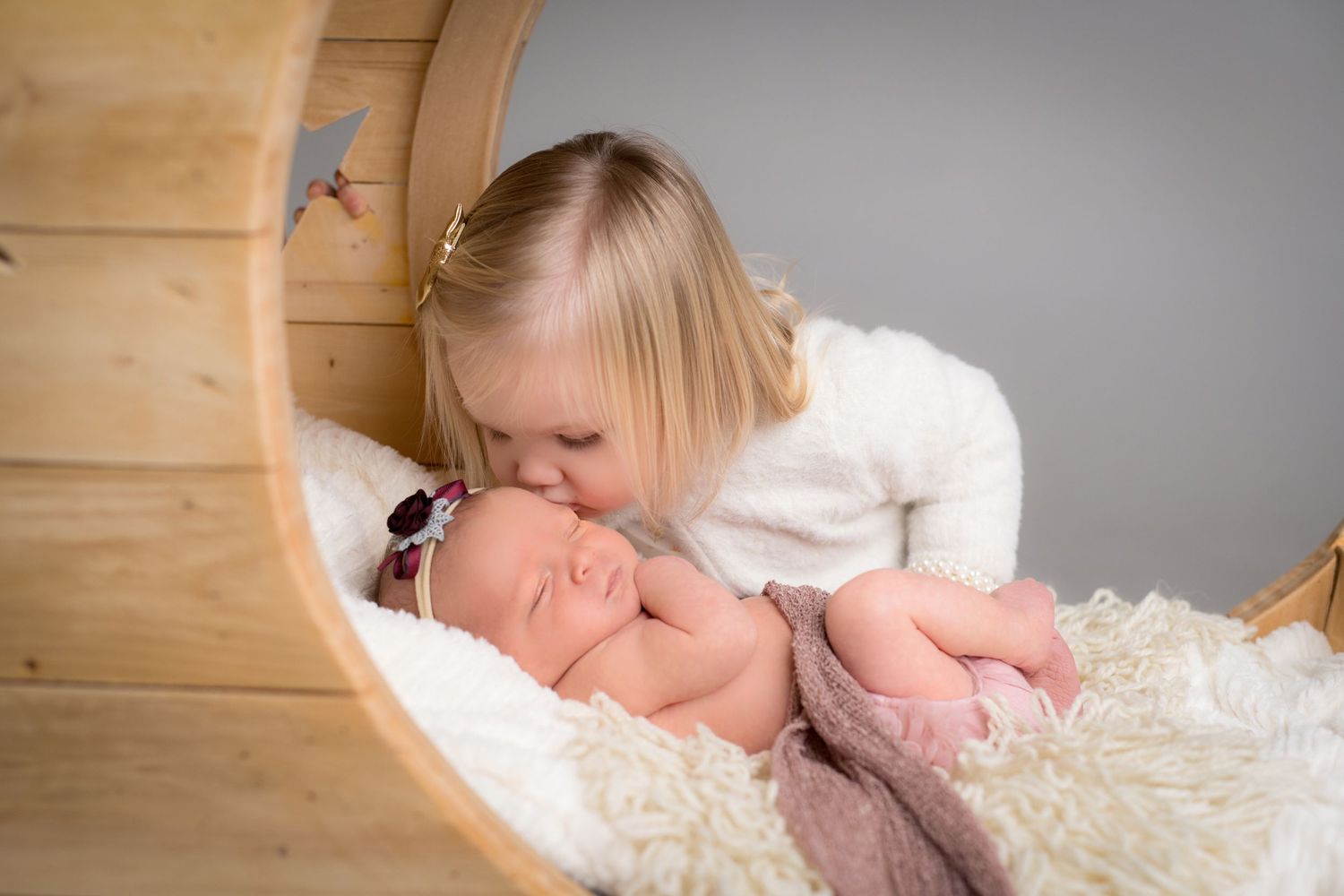 Baby sleeping on white fluffy blanket in a wooden cradle while being kissed by sibling.